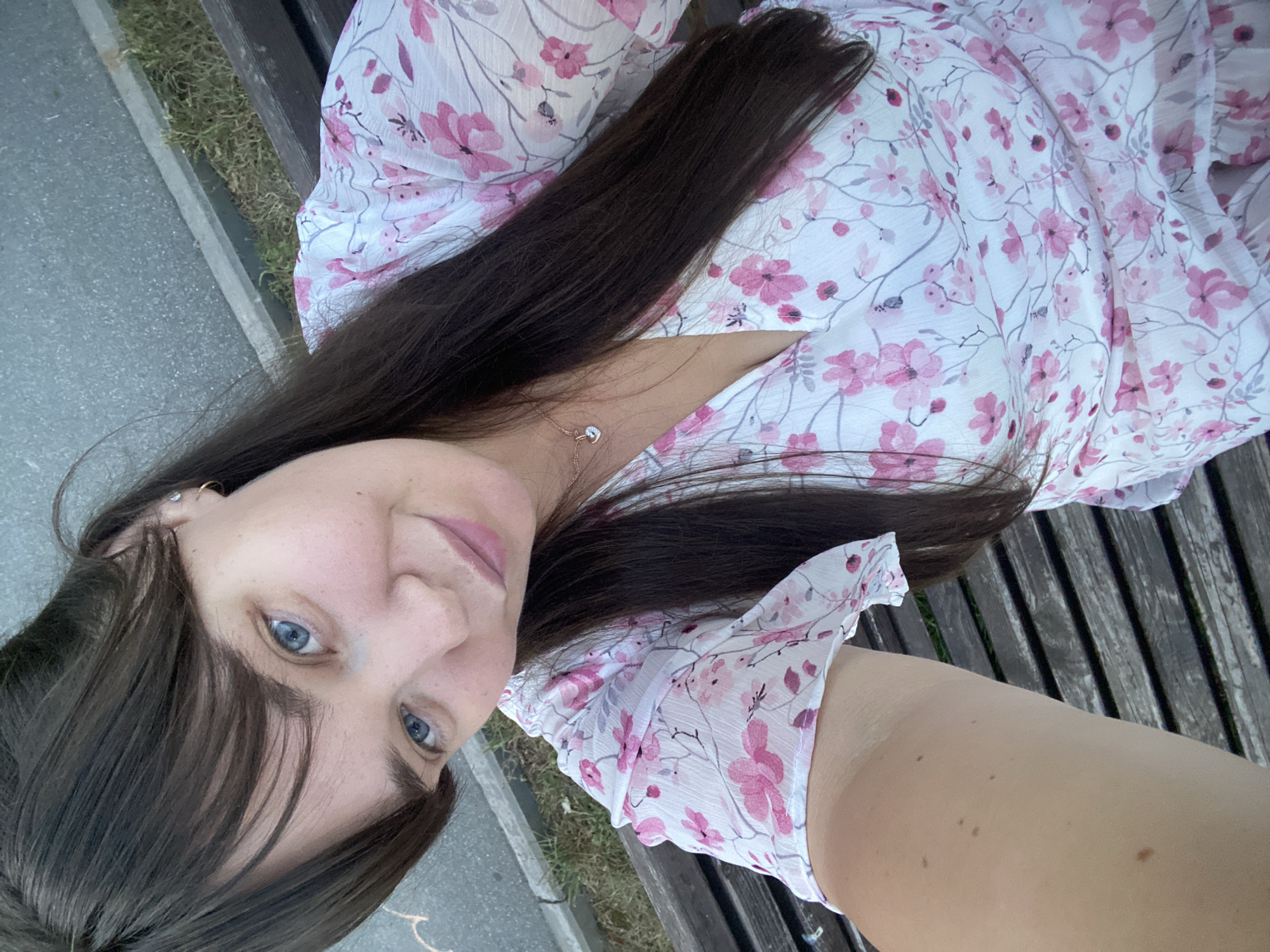 A young woman with long, dark hair and blue eyes takes a selfie on a park bench. She is wearing a white dress with a floral pattern of pink flowers and green leaves. She has a small silver necklace with a heart-shaped pendant. The bench is made of dark wood and is located outdoors, likely in a park or public space, with a paved walkway and grass visible in the background. The lighting suggests it is daytime. The image is taken from a high angle, looking down at the woman.