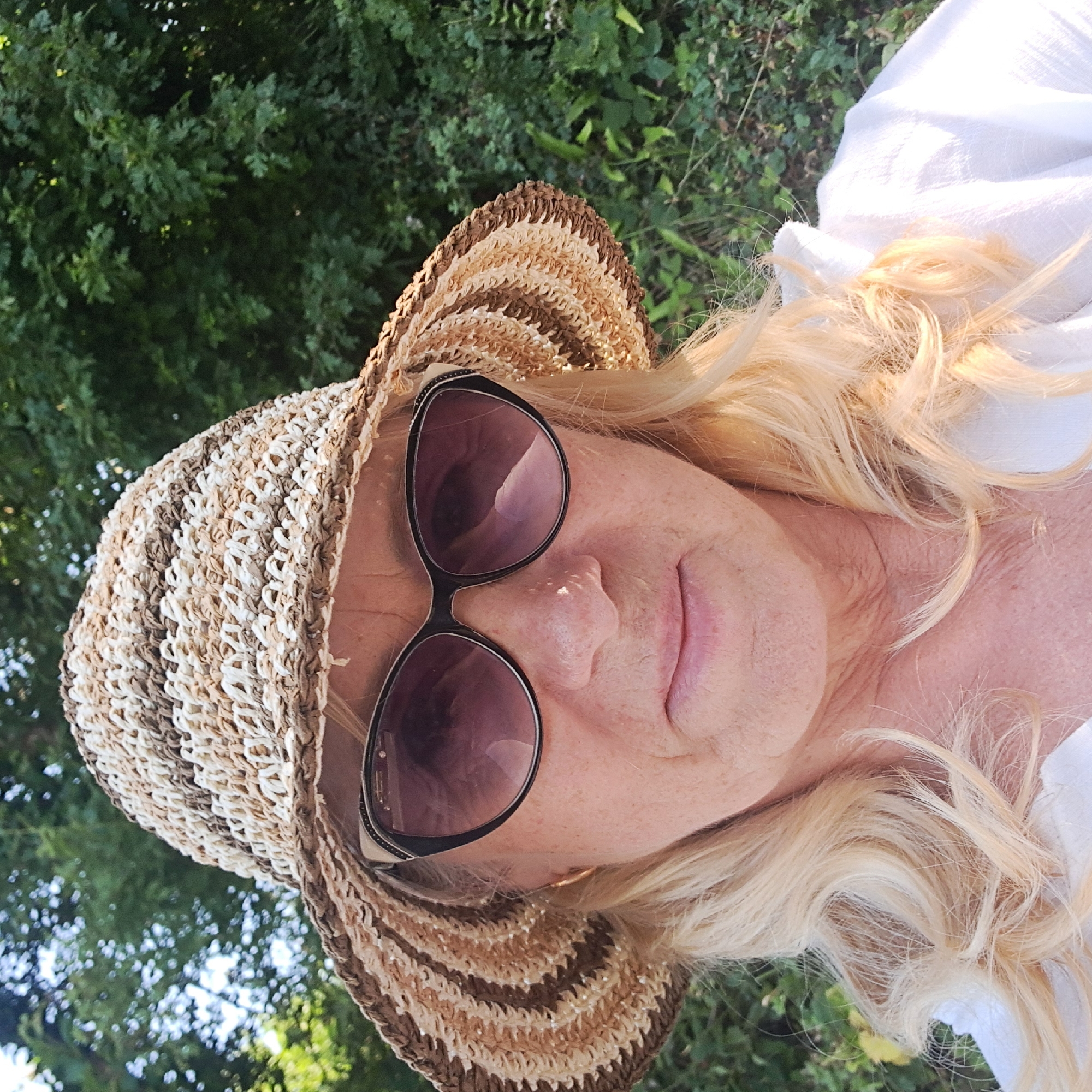 A close-up, waist-up shot features a Caucasian woman with blonde, wavy hair, wearing a wide-brimmed straw hat with alternating brown and white stripes, and dark-lensed sunglasses. She is dressed in a white collared shirt. The background consists of blurred green foliage, suggesting an outdoor setting, possibly a garden or park. The lighting indicates it is daytime. There are no other individuals present in the frame. The image appears to be taken outdoors in a natural, green environment, possibly in Ireland, though no specific city or landmark is identifiable from the visual information alone.