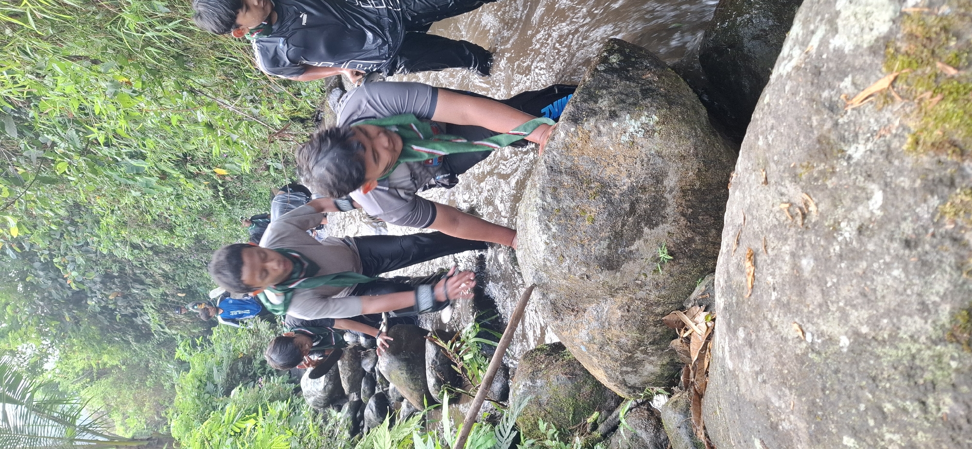In the city of Puntukrejo, Indonesia, a group of young individuals, likely scouts given their attire and scarves, are navigating a shallow, rocky stream surrounded by lush green vegetation. The water is clear, revealing the submerged rocks and the movement of the stream. The individuals are wearing dark-colored athletic clothing, with some sporting green and white patterned scarves around their necks. They are in various stages of crossing the stream, some are climbing over large rocks while others are wading through the water. One individual in the foreground is captured mid-climb, bracing themselves against a large, moss-covered boulder. The environment is dense with foliage, including palm leaves and other broad-leafed plants, indicating a natural, possibly forested, setting. The lighting suggests an overcast day or a shaded area, as there are no strong shadows. The overall scene depicts an outdoor activity, likely an adventure or training exercise, in a natural stream environment.