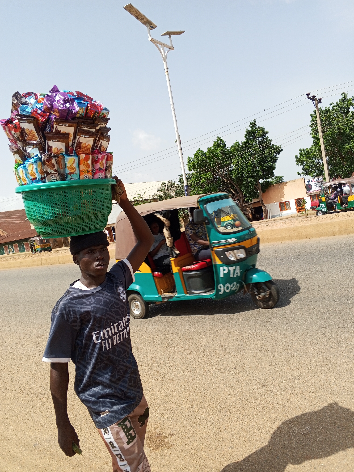 The image shows a young male vendor in Bukuru, Nigeria, carrying a large green basket filled with packaged snacks on his head. He is wearing a dark blue t-shirt with white lettering that reads "Emirates Fly Better" and patterned trousers. The vendor is walking on a paved road under a clear blue sky. In the background, a teal and yellow auto-rickshaw with the license plate "PTA 9029" is visible, along with other auto-rickshaws and buildings lining the street. Trees and power lines are also present in the scene. The overall impression is of a busy street scene in an African city.