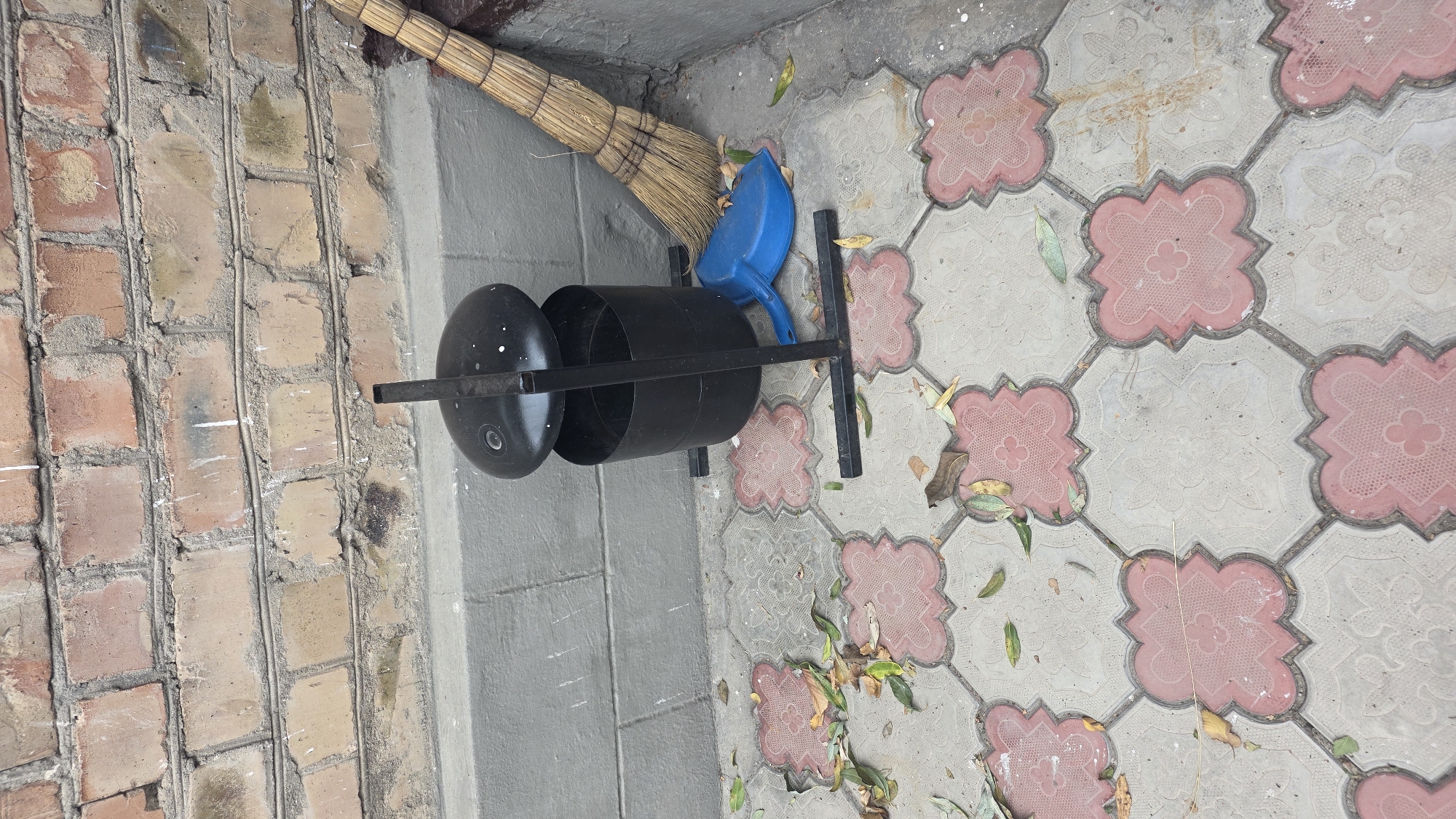 The image depicts an outdoor scene on a paved area with decorative tiles, likely in an urban setting in Tokmok City, Kyrgyzstan. In the background, a brick wall shows signs of aging and some white paint splatters. A gray concrete ledge runs along the base of the wall.
The primary subjects in the foreground are a black metal trash receptacle with an open lid, a blue plastic dustpan, and a broom with straw bristles leaning against the trash receptacle. The trash can is supported by a black metal stand with four legs. The dustpan is positioned on the ground next to the trash can. The broom is placed in the corner, leaning against the wall and trash receptacle.
The paved ground is covered with interlocking tiles. Some of these tiles are light gray, while others are a reddish-pink color with a decorative four-petal flower motif. Scattered across the paved area are several fallen leaves, indicating an outdoor environment, possibly in autumn.
The scene appears to be set up for outdoor cleaning or maintenance, with the broom and dustpan ready for use. There are no people visible in the image. The lighting suggests it is daytime, with no strong shadows indicating direct sunlight.