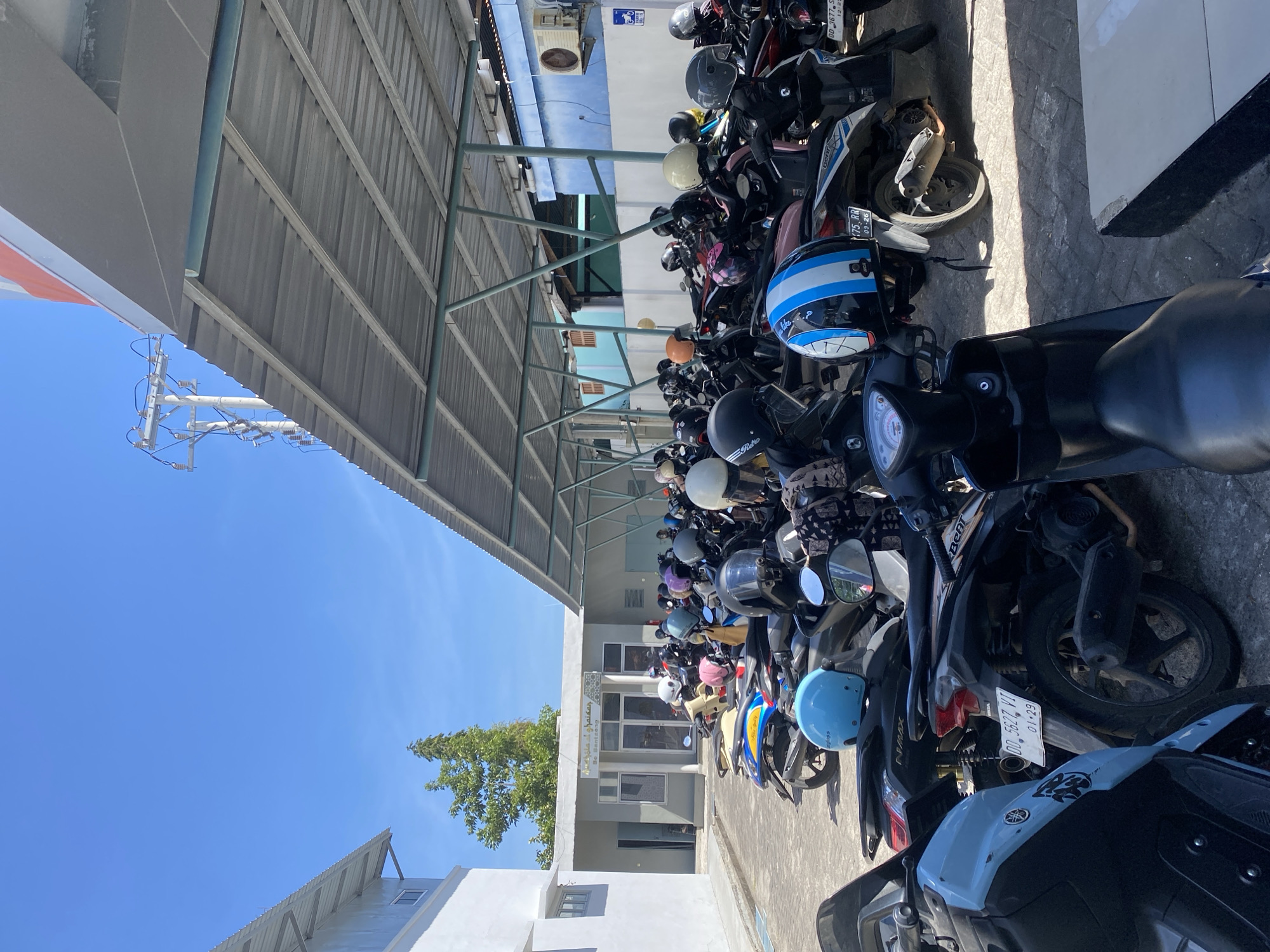 The image shows a parking lot filled with motorcycles under a covered structure. The lot is paved with gray interlocking bricks. In the background, a light blue building with white trim has a sign above its entrance that reads "Mc. Santeng" in Arabic script and Latin script. To the left of the building, a tree with green foliage stands against a clear blue sky. A utility pole with electrical wires is visible in the distance. Several motorcycles, mostly scooters, are parked in rows under the corrugated metal roof of the shelter. Many of the motorcycles have helmets on them. The overall scene suggests a public area where people park their vehicles, potentially a school, mosque, or community center in Kelurahan Tappanjeng, Indonesia.