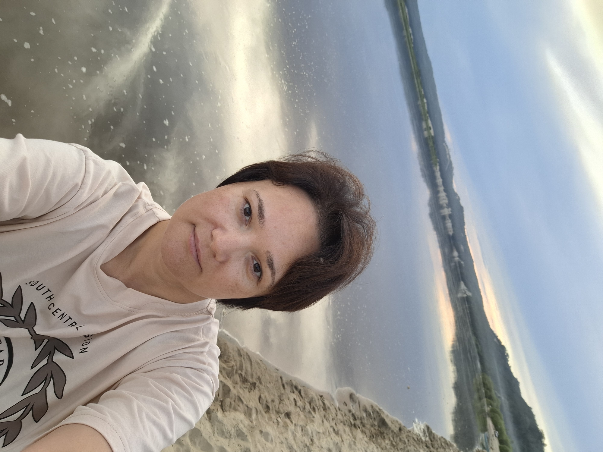 In the city of Malaya Sazanka, Russia, a woman of Asian descent with short, dark brown hair and fair skin with freckles is taking a selfie on a sandy beach next to a large body of water. She is wearing a light-colored t-shirt with a graphic print that reads "SOUTH CENTRAL UNION." The water reflects the sky, which is filled with a mixture of blue and white clouds, with a hint of orange and yellow on the horizon, suggesting either sunrise or sunset. In the background, a tree-lined hillside rises from the water. The overall scene is serene and appears to be a natural landscape.