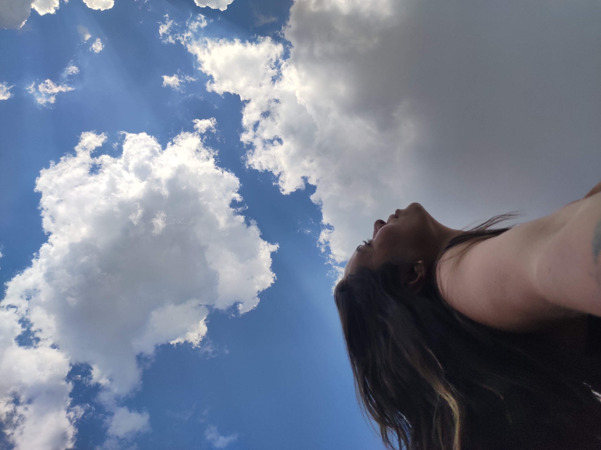 The image depicts a low-angle shot of a person looking upwards at the sky. The sky is partially cloudy, with some areas of clear blue sky and other areas covered by white, fluffy clouds. Sunlight appears to be shining through the clouds. The person's face and torso are visible in the bottom and right of the frame, with long brown hair cascading down. The location of the photo is not specified, but the prompt stated that the city is in Фоки, Russia.