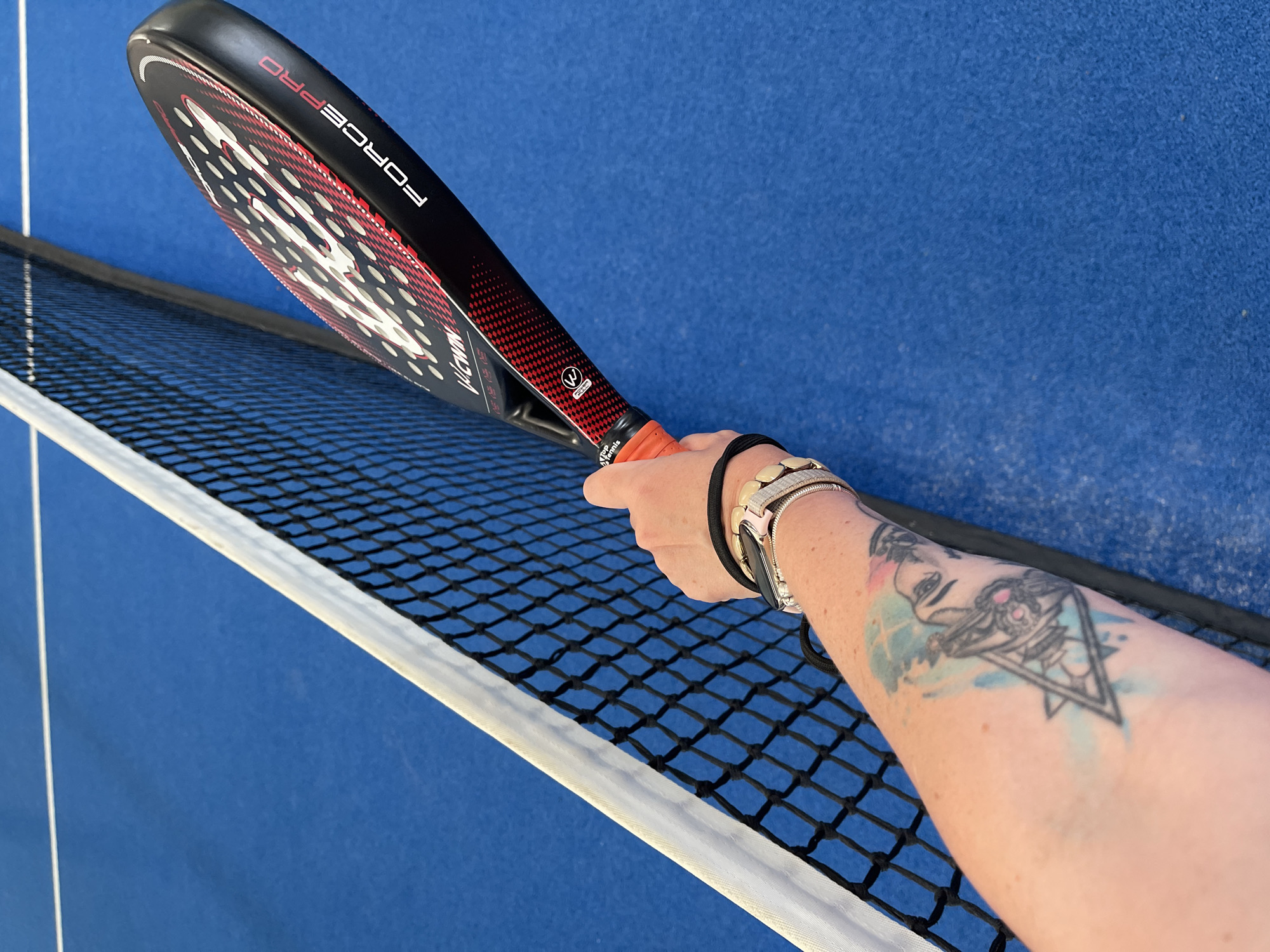 An arm with tattoos and a watch is holding a black and red padel racket over a blue padel court near a net. The racket has "FORCE PRO" printed on it. The net is black with a white border. The blue court has a white line across it.