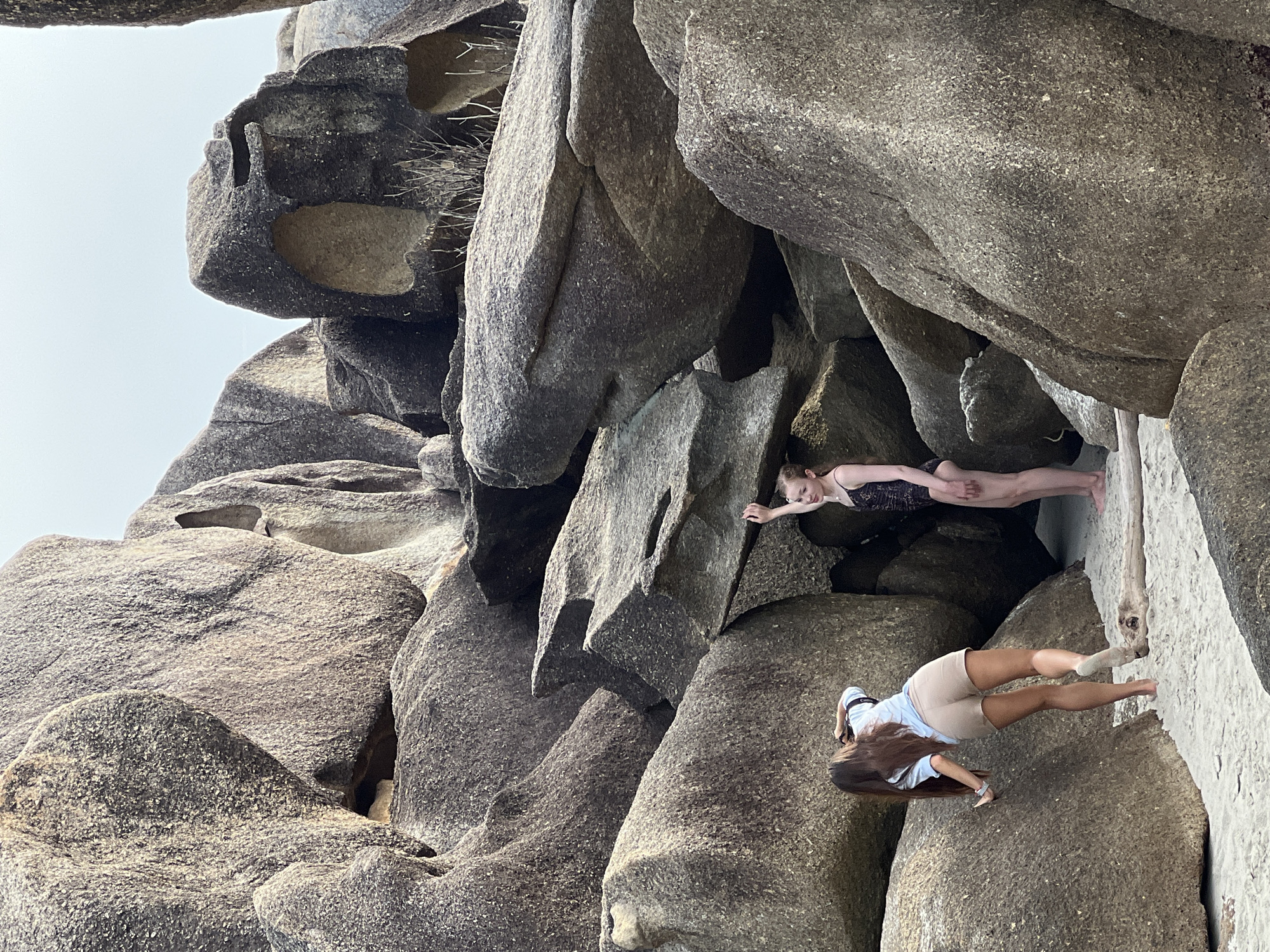 Two young women are on a sandy beach surrounded by large, weathered granite boulders. The woman on the left, with long dark hair, is wearing a light blue shirt and beige shorts and is bent over, looking down and touching a boulder. Her right leg is extended behind her. The woman on the right is wearing a dark patterned swimsuit and is standing, with her left arm raised and her hand touching a boulder above her. She is looking towards the left. The boulders are characterized by smooth, rounded surfaces and some have hollowed-out areas. There is a piece of driftwood on the sand between the two women. The sky is overcast. The location is identified as Baan Chaweng Noi, Thailand.