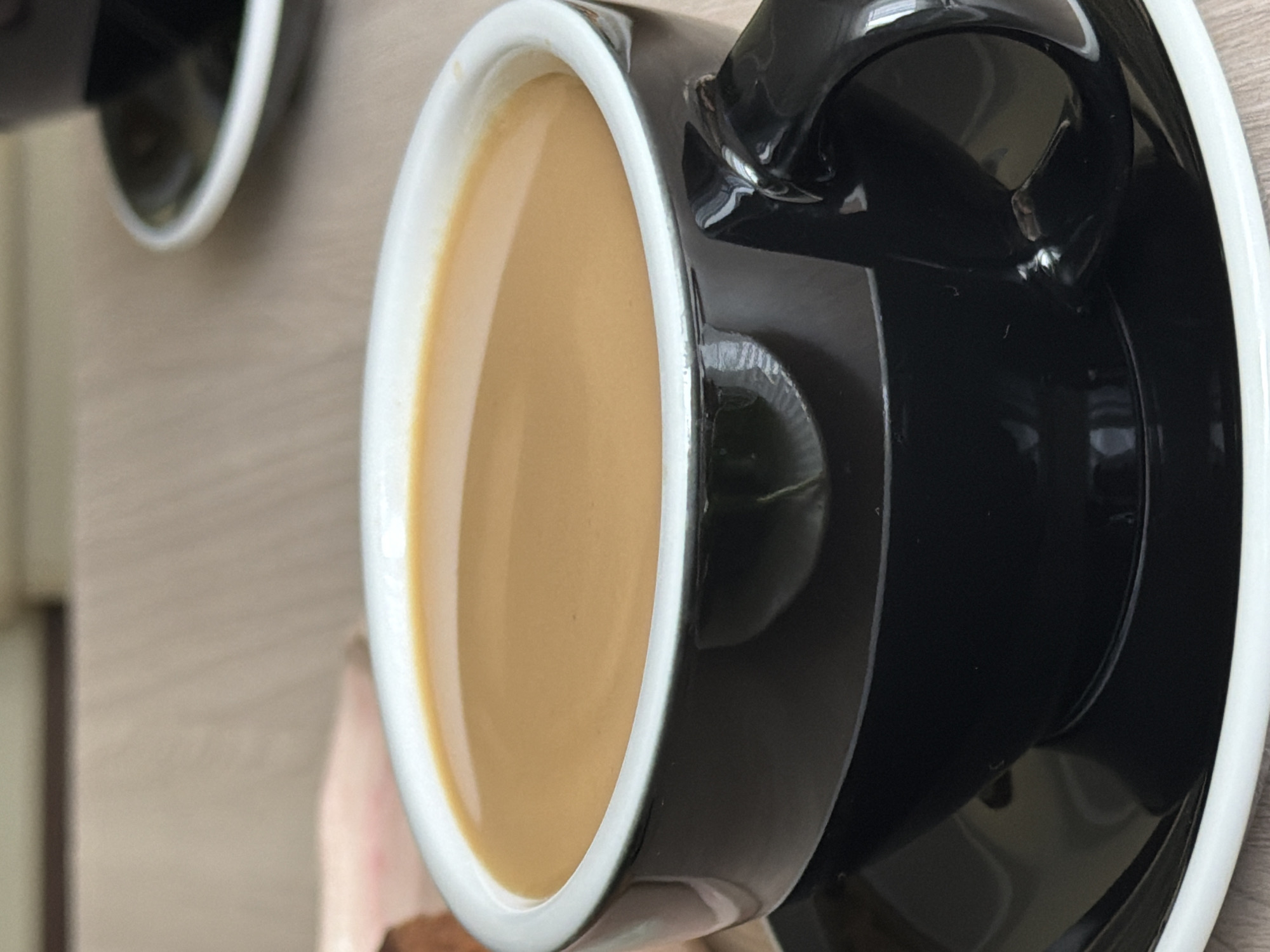 A close-up shot shows a black coffee cup filled with a light brown beverage, likely coffee with milk or cream. The cup has a thick white rim and sits on a matching saucer with a white rim. A second cup is partially visible in the upper left corner, suggesting a pair. The surface underneath is a light brown, possibly a wooden table. The scene appears to be indoors, possibly in a cafe or home setting.