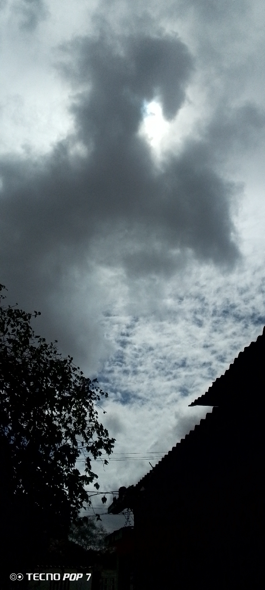 The image shows a cloudy sky above a silhouetted roofline and tree foliage. The sun is partially visible through a gap in the dark, heavy clouds, creating a bright, blown-out area in the upper center of the frame. Below the sky, the dark silhouette of a tiled roof slopes downwards from the right side of the image. To the left, the silhouette of a tree with leaves and branches extends into the frame from the left edge. Power lines are visible stretched horizontally between the tree and the roofline, with some faint details of buildings and other structures visible in the darker shadows below the roof. The bottom of the image contains a watermark reading "TECNO POP 7". The scene appears to be an outdoor view, possibly from a residential area, under overcast conditions. The location is identified as Altagracia, Venezuela.