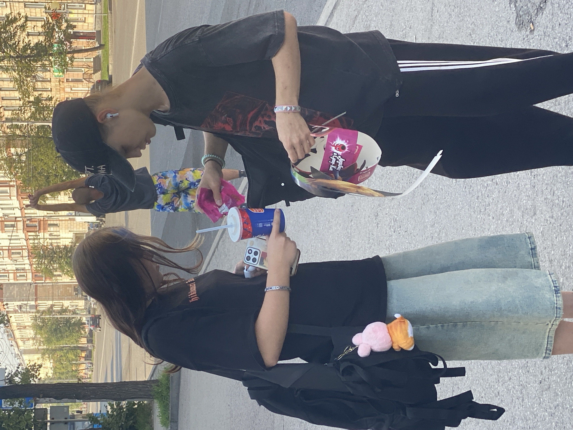 Two young individuals, likely teenagers, are standing on a sidewalk in an urban environment. The person on the left, with long brown hair and a black t-shirt, is holding a smartphone and a beverage cup. A black backpack is slung over their shoulder, adorned with a pink stuffed animal keychain. They are wearing a light blue denim skirt. The person on the right, wearing a black t-shirt and a black baseball cap, is holding a piece of paper with graphics. They have white earbuds in and are wearing black sweatpants with white stripes. A third person is visible in the background, stretching with their arms raised, wearing a dark t-shirt and colorful shorts. The setting appears to be a street with buildings and trees, suggesting a public area in a city. The overall scene captures a casual moment between the two individuals, possibly during an outing. The presence of street signs and architectural styles of the buildings could aid in pinpointing the exact location within Novokuznetsk, Russia.