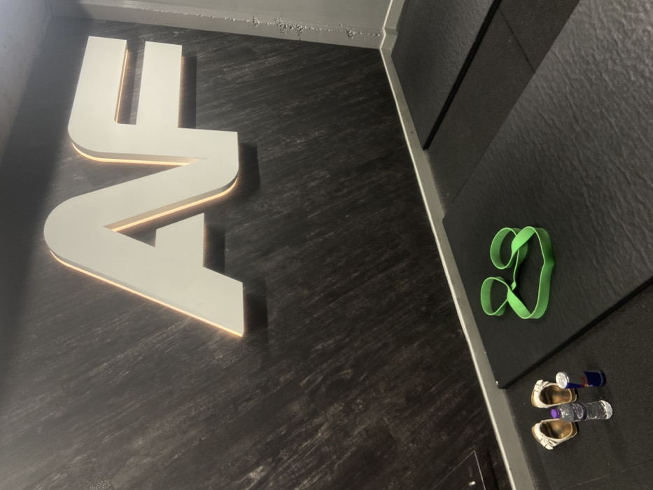 The scene depicts an interior view of what is identifiable as an Anytime Fitness gym, likely located in London, United Kingdom. A large, white, backlit "AF" logo is prominently displayed on a dark, textured wall. The floor is covered with black, textured exercise mats. On the leftmost mat, a green resistance band is loosely arranged. Adjacent to this mat, on the floor, are a pair of light-colored sandals with gold-toned buckles, a small plastic water bottle with a blue cap, and a red Red Bull energy drink can. No individuals are present in the frame.