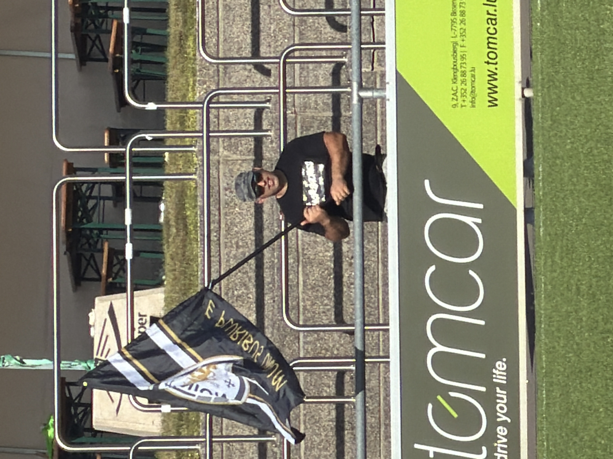 A medium shot, taken in daylight, presents a man standing behind a silver-toned railing, seemingly at a sporting event, given the green patch resembling a field. The man is dark-haired, wearing a cap and sunglasses, and a dark t-shirt adorned with a graphic. He is partially obscured by the railing, but is positioned next to a black, gold, and white flag.
To the right of the man is a large sign advertising "tomcar" with the tagline "drive your life." The sign includes business contact information in Luxembourg (9, ZAC Klengbousbierg 1-7795 Bissen; T+352 26887395; info@tomcar.lu; www.tomcar.lu). The background shows outdoor seating, including green benches and wooden tables. The setting suggests an outdoor venue possibly in Fentange, Luxembourg, based on the address listed.
