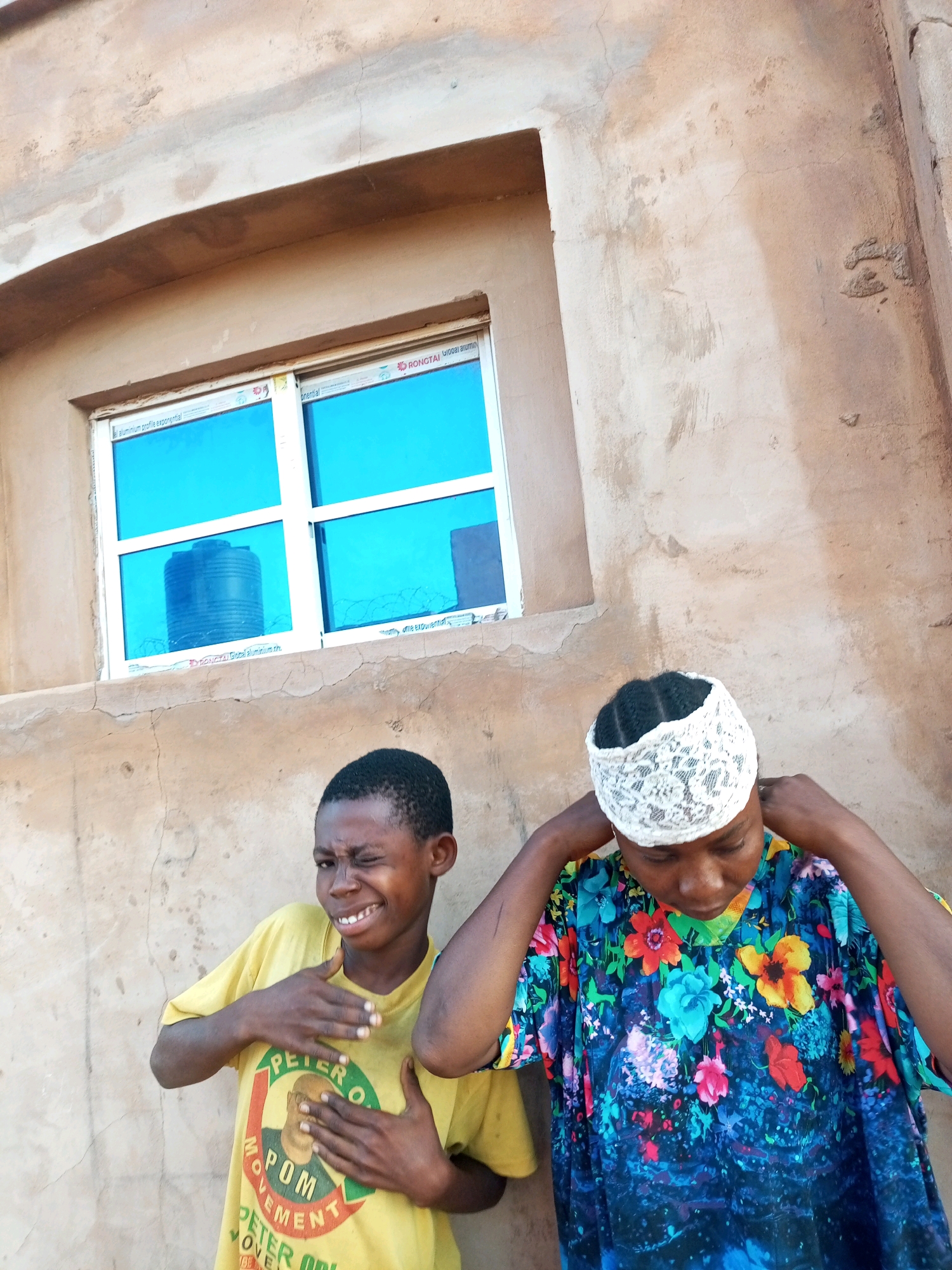 The image shows two individuals standing in front of a building in Tempure, Nigeria. On the left, a young boy is wearing a yellow t-shirt with a circular emblem that reads "PETER O." and "MOVEMENT" with a portrait of a man in the center. He is winking and has his hands placed on his chest and arm. To the right, a person is wearing a white lace headscarf and a colorful floral-patterned shirt. They are looking down and adjusting their headscarf. Behind them is a building with a textured, beige-colored wall and a window with bright blue panes reflecting an outdoor scene. A large black water tank is visible through the window. The environment appears to be outdoors, with the building's rough stucco wall indicating a potentially arid or developing urban setting.