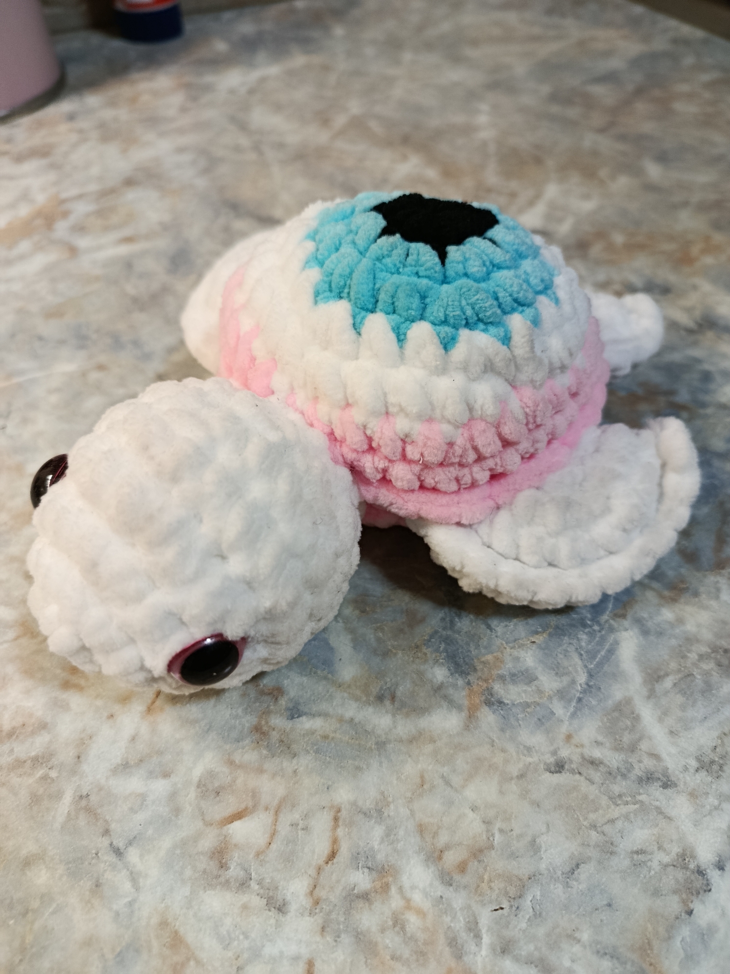 A crocheted turtle toy with a white head and limbs, and a multi-colored shell featuring white, pink, and light blue stripes, and a black center, is placed on a marble-patterned surface. The turtle's head is facing the left, with two visible black button eyes. The background is out of focus, showing indistinct objects on the left side. The scene does not depict any identifiable location within the Municipal Unit of Rhodes, Greece.