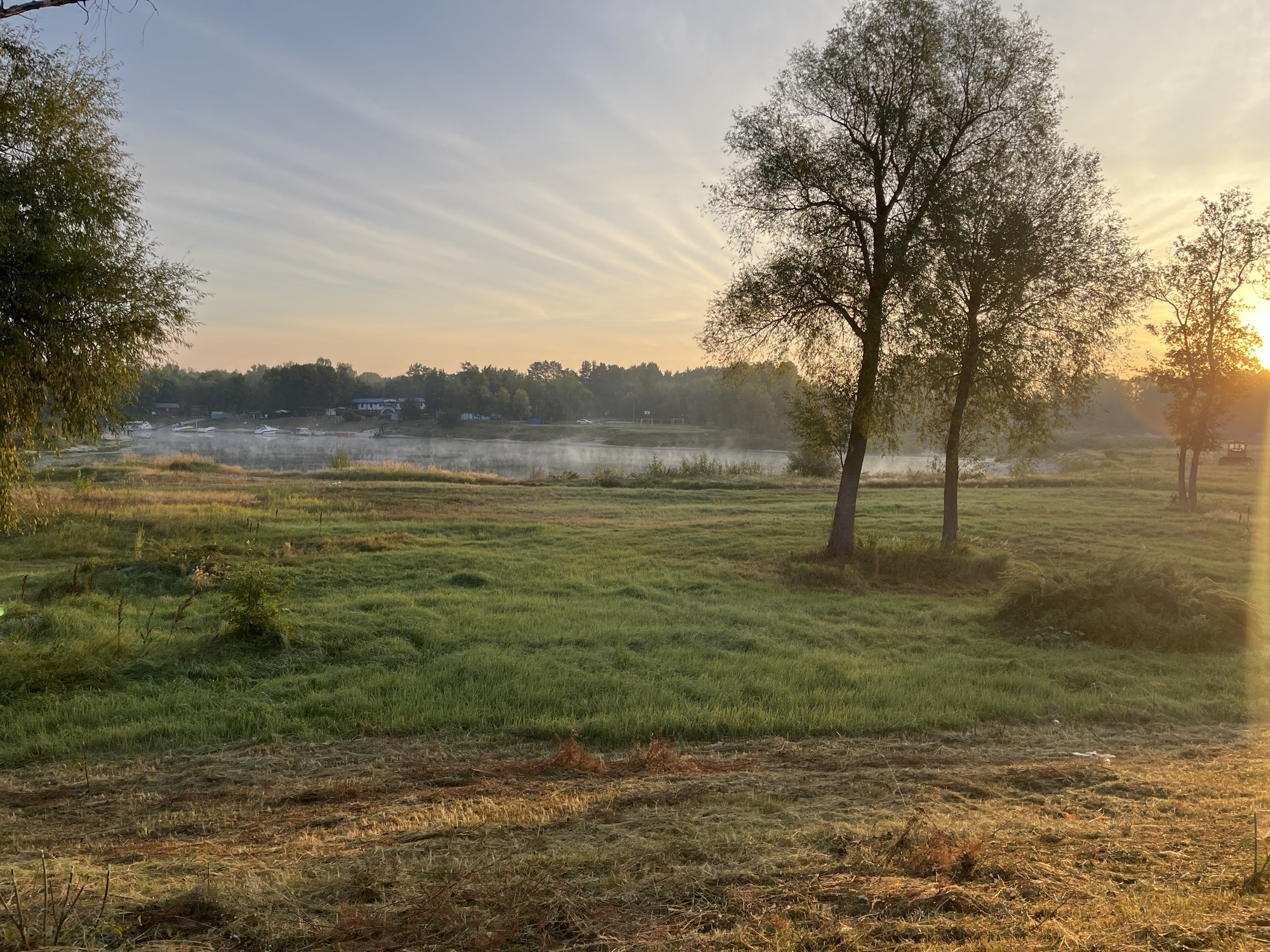 This is a scenic landscape photograph taken at sunrise over a body of water, likely a river or lake. In the foreground, a grassy field with dew or mist is illuminated by the low-lying sun. Several trees, with their branches silhouetted against the sky, are scattered across the scene. In the middle ground, the water is visible with a light mist rising from its surface. Along the far bank of the water, there is a treeline with some buildings and a few boats docked. The sky is clear with wispy clouds that radiate from the sun, creating a dramatic effect. The overall impression is one of tranquility and natural beauty, typical of a rural or semi-rural setting.