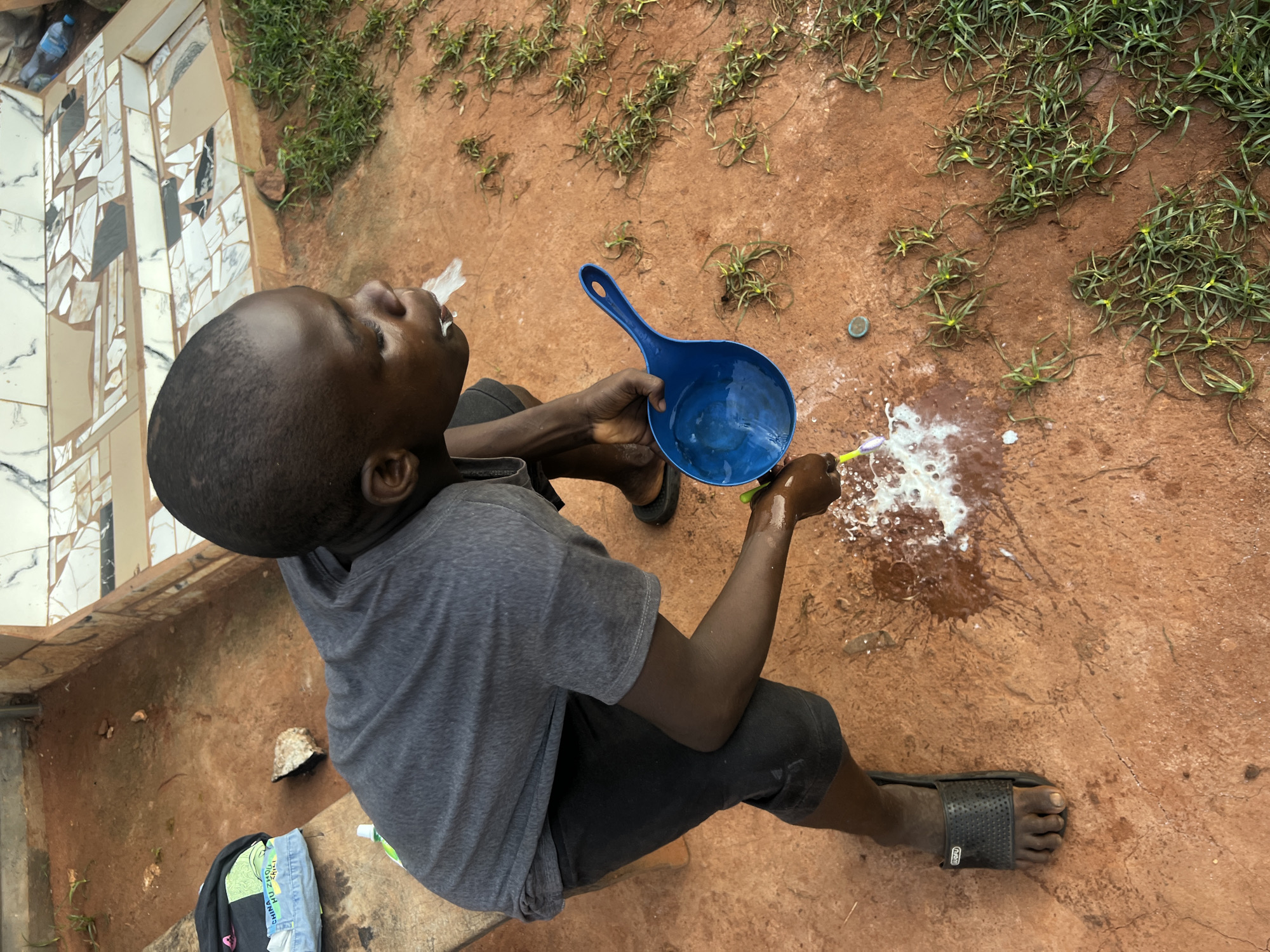 The image depicts a young male outdoors in Obosi, Nigeria. He is seated on a reddish-brown, cracked earth surface with sparse green vegetation. The individual is wearing a gray t-shirt, black shorts, and black sandals. He is rinsing his mouth with water from a blue plastic scoop, spitting water and toothpaste onto the ground. A toothbrush is held in his right hand. Behind him is a wall with decorative tiles. A bag with clothing is near the wall.
