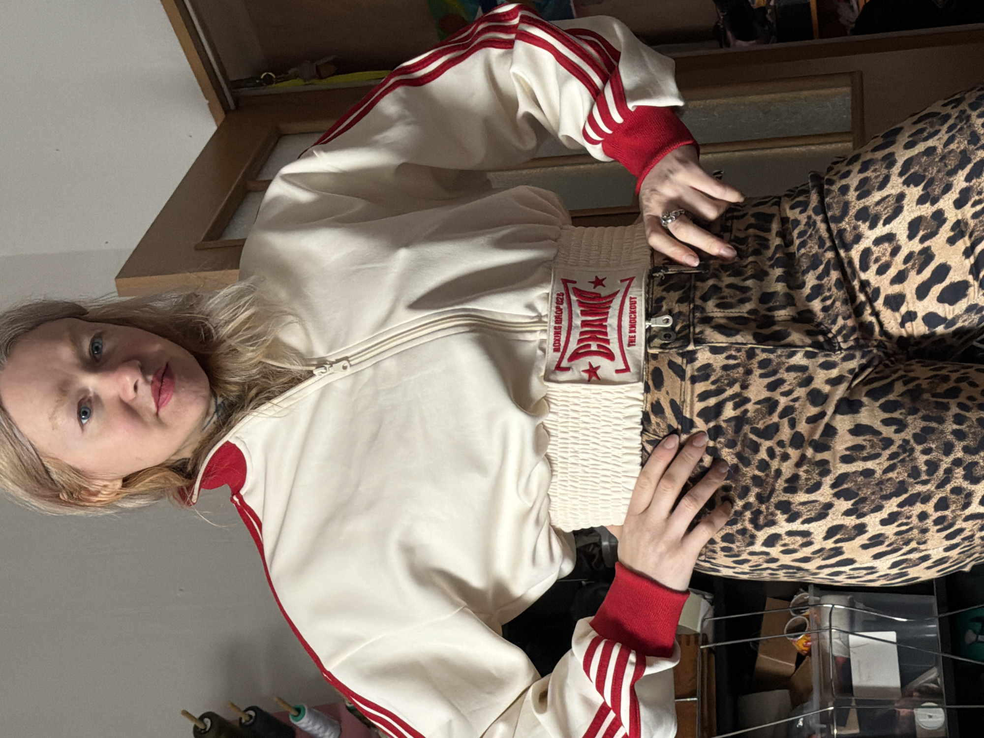 An adult female individual is depicted standing indoors in what appears to be a residential room in Saint Petersburg, Russia. The subject has fair skin, light-colored hair, and blue eyes, with a neutral facial expression while looking directly at the camera. She is wearing a cream-colored, cropped track jacket with a zipper closure, red collar, red cuffs, and three red stripes extending down each sleeve. The jacket's elasticized waist features a white rectangular patch with red embroidery, reading "BOXING SHOP 025," a large "CHAMP" with two stars, and "THE KNOCKOUT." Below the jacket, she wears leopard-print trousers. Her hands are positioned on her hips or upper thighs, with a ring visible on her right ring finger. The background consists of a plain light-colored wall to the left and a brown wooden door or closet door to the right. On the left side of the frame, spools of thread on wooden spindles and a clear plastic storage container are visible on a shelving unit.