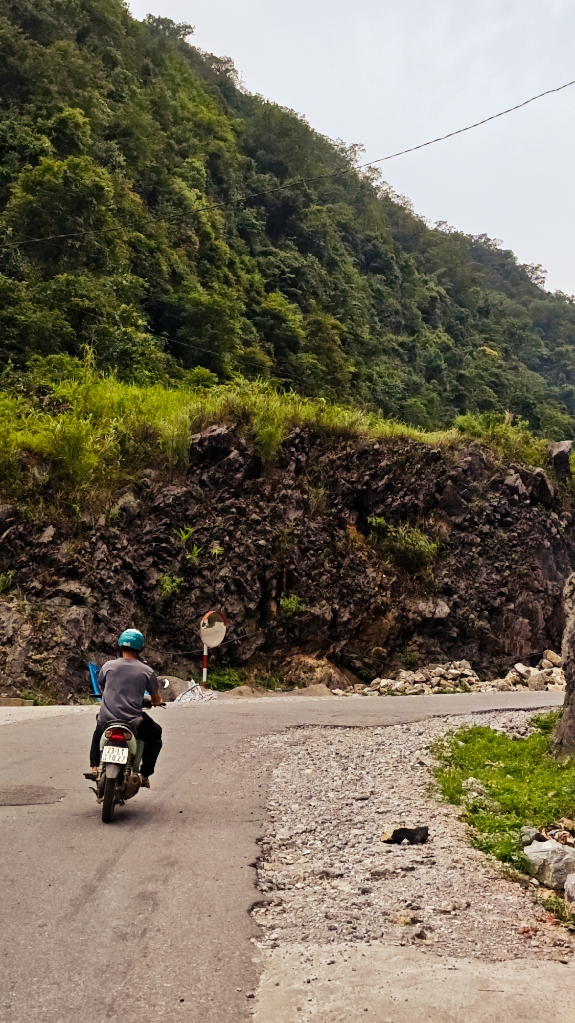 A motorcyclist with a blue helmet is traveling on a paved road in the Cán Tỷ Commune, Vietnam. The road is surrounded by a steep, rocky hillside covered in lush, green vegetation. A convex mirror is mounted on a pole along the roadside. The road surface appears to be patched with gravel and is in a state of disrepair.