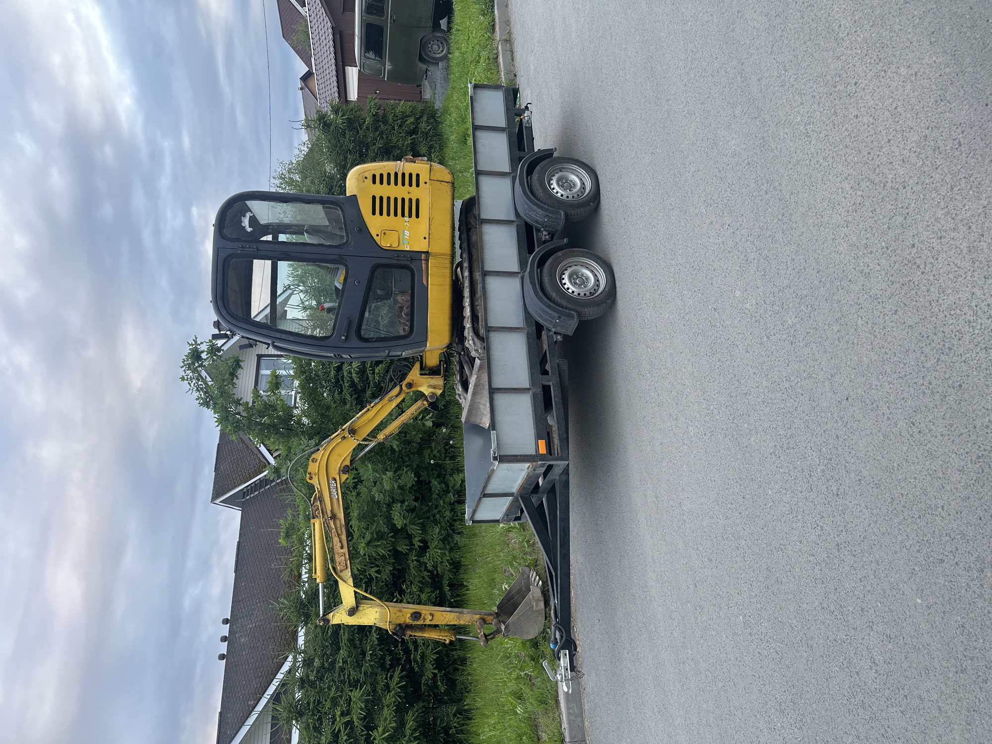 A yellow mini excavator is loaded onto a grey, tandem-axle trailer. The trailer is positioned on an asphalt road adjacent to a curb and a grassy verge. Behind the verge, dense green bushes form a hedge, with houses and trees visible beyond. The sky is overcast with grey clouds. The scene appears to be in a residential area of Petrozavodsk, Russia.