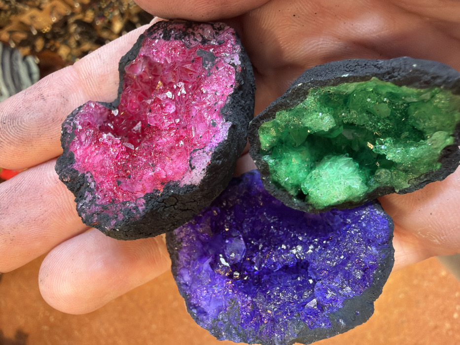A close-up image taken in Arucas, Spain, depicts an individual's hand holding three distinct, colorful geode formations. The hand, exhibiting visible skin texture and minor dark smudges on the fingers, serves as the primary support for the objects. The uppermost geode presents a vibrant fuchsia or hot pink crystalline interior, encased by a rough, dark, charcoal-like exterior shell. To the lower left, a second geode displays a brilliant purple crystalline structure within a similar dark shell. The third geode, situated to the lower right, features vivid green crystals also contained within a dark, rugged outer layer. All three geodes exhibit crystalline facets that reflect light. The background is blurred, showing warm, earthy tones consistent with an indoor environment, possibly a wooden surface. No other individuals or specific actions are discernible beyond the hand holding the objects.