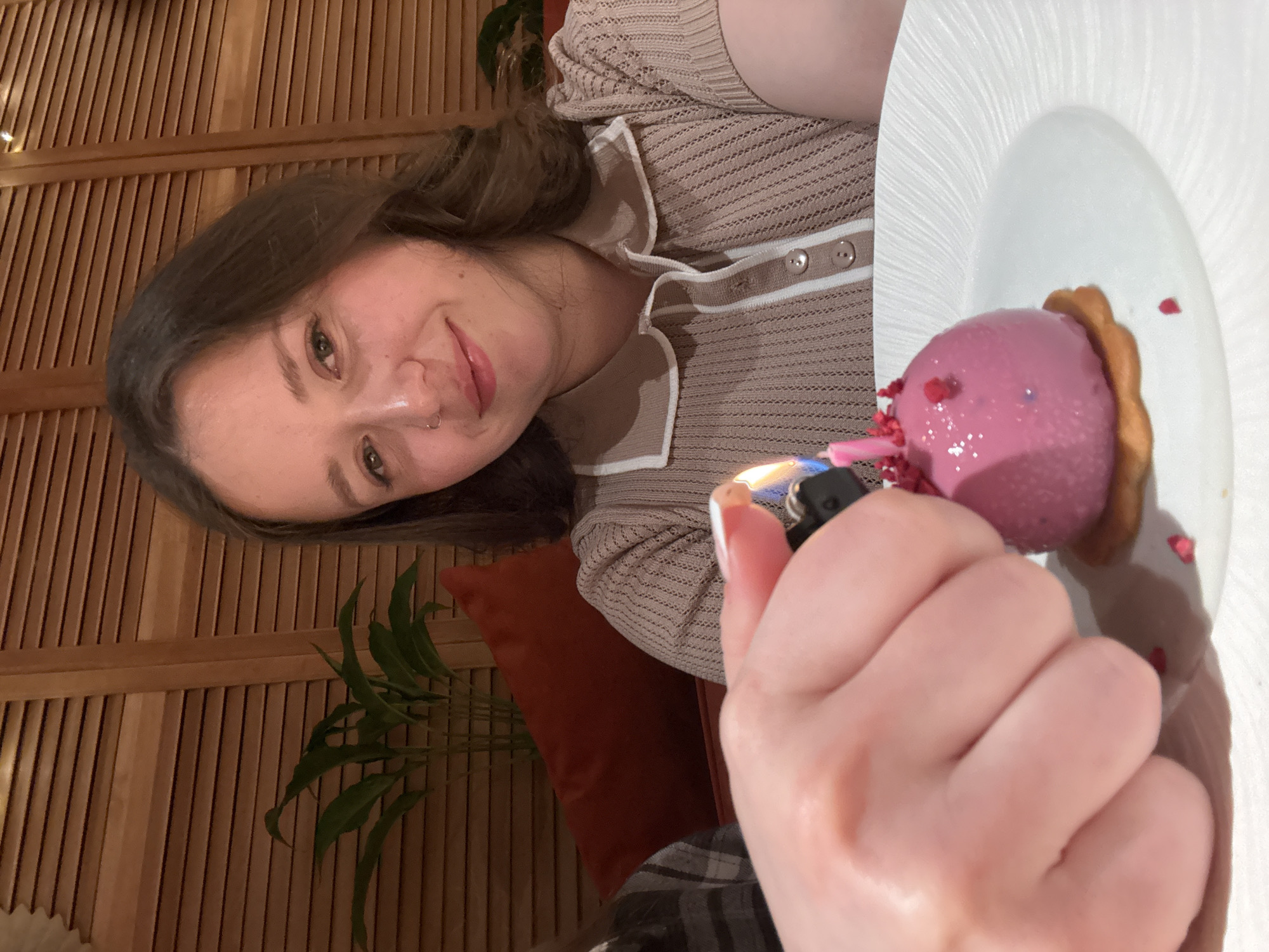 An indoor, close-up shot captures a young woman lighting a candle on a dessert. The woman, with long brown hair, fair skin, and light eyes, looks towards the camera with a gentle smile. She has a subtle nose piercing and wears a light brown, possibly beige, knitted top with a striped collar and buttons down the front.
In the foreground, a hand, likely hers, holds a black lighter with a visible flame, igniting a small pink stick candle embedded in a spherical pink dessert. The dessert, possibly a mousse or ice cream, is garnished with tiny red sprinkles or dried fruit pieces and a small, fluted, light brown cookie or pastry. It rests on a white, textured plate.
The background features warm-toned wooden slatted panels, suggesting a wall or room divider in a restaurant or cafe. A glimpse of a dark green plant and an orange/rust-colored cushion are visible, adding to the cozy ambiance. The lighting appears soft and intimate, indicative of an evening setting. The overall scene suggests a celebratory moment, possibly a birthday. No text is visible in the image. The location is Izhevsk, Russia.