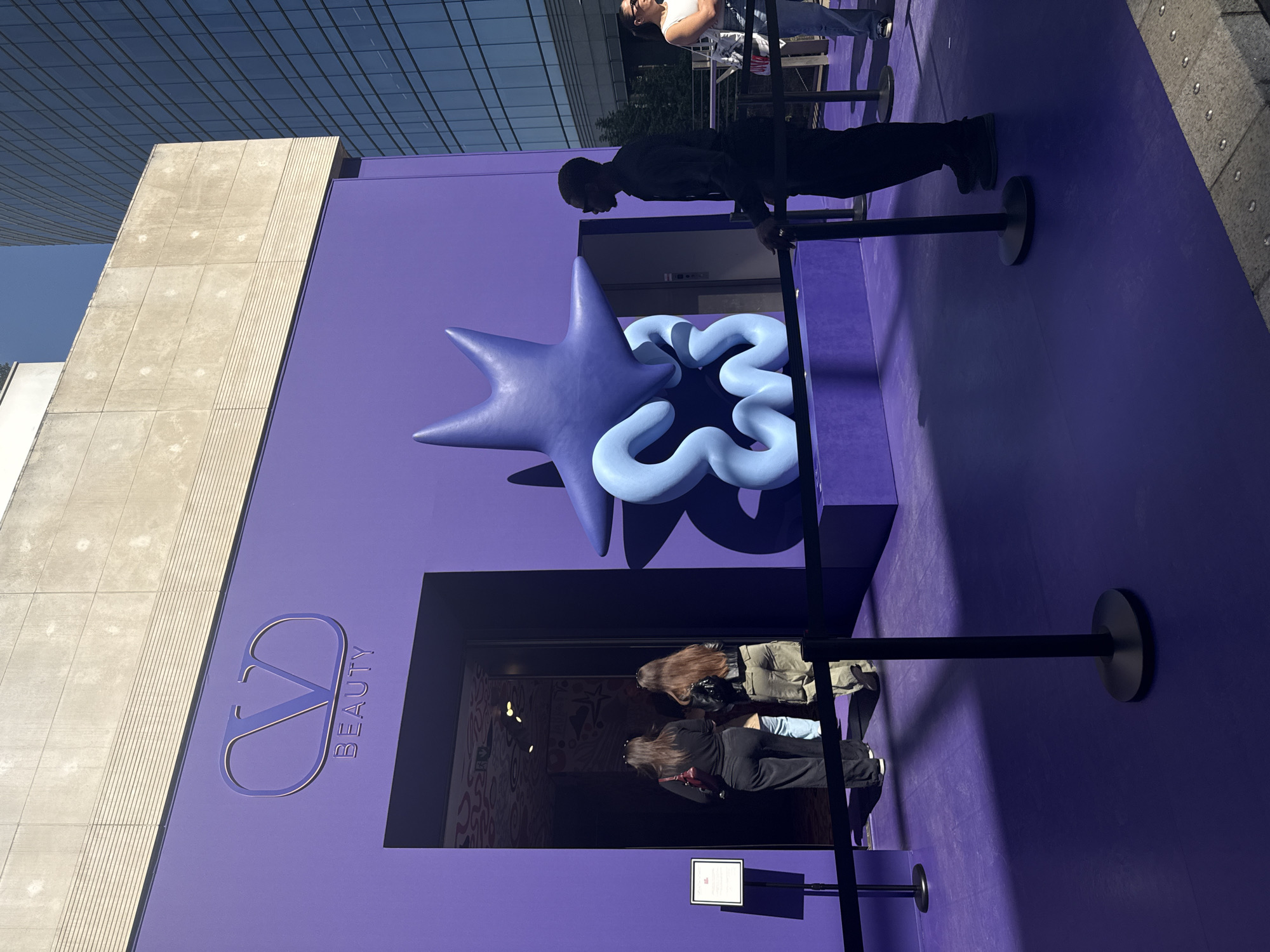 The image captures a vibrant, outdoor scene featuring a temporary pop-up store or event entrance for Valentino Beauty, set against an urban backdrop under bright sunlight. The dominant element is a large, striking purple structure, which serves as the facade of the establishment.
Protruding from the purple wall are two large, sculptural objects: a prominent, dark blue, multi-pointed star-like form, and a lighter blue, organic, squiggly shape nestled behind it. Below these sculptures, an open entrance reveals a dark interior with some visible wall decorations.
Several people are present. A person dressed in black stands to the right, appearing to queue or attend to the entrance. Two individuals, viewed from behind, are positioned directly in front of the doorway, peering into the interior. Further in the background to the right, another person wearing a white top and sunglasses is visible. Black stanchions with connecting ropes are set up, guiding potential visitors into a queue.
On the left side of the purple structure, the logo "VΛ BEAUTY" (where 'V' and 'Λ' are stylized to form the brand's 'VD' monogram) is embossed in a slightly lighter shade of purple, clearly identifying the brand.
The background shows modern architecture, including a light-colored concrete building and the reflective glass facade of a high-rise, suggesting a contemporary city setting. The bright sunlight casts strong shadows on the purple ground and surrounding areas, indicating a clear, sunny day. The overall impression is that of a modern, stylish brand activation in a lively urban location, such as Milan, Italy.