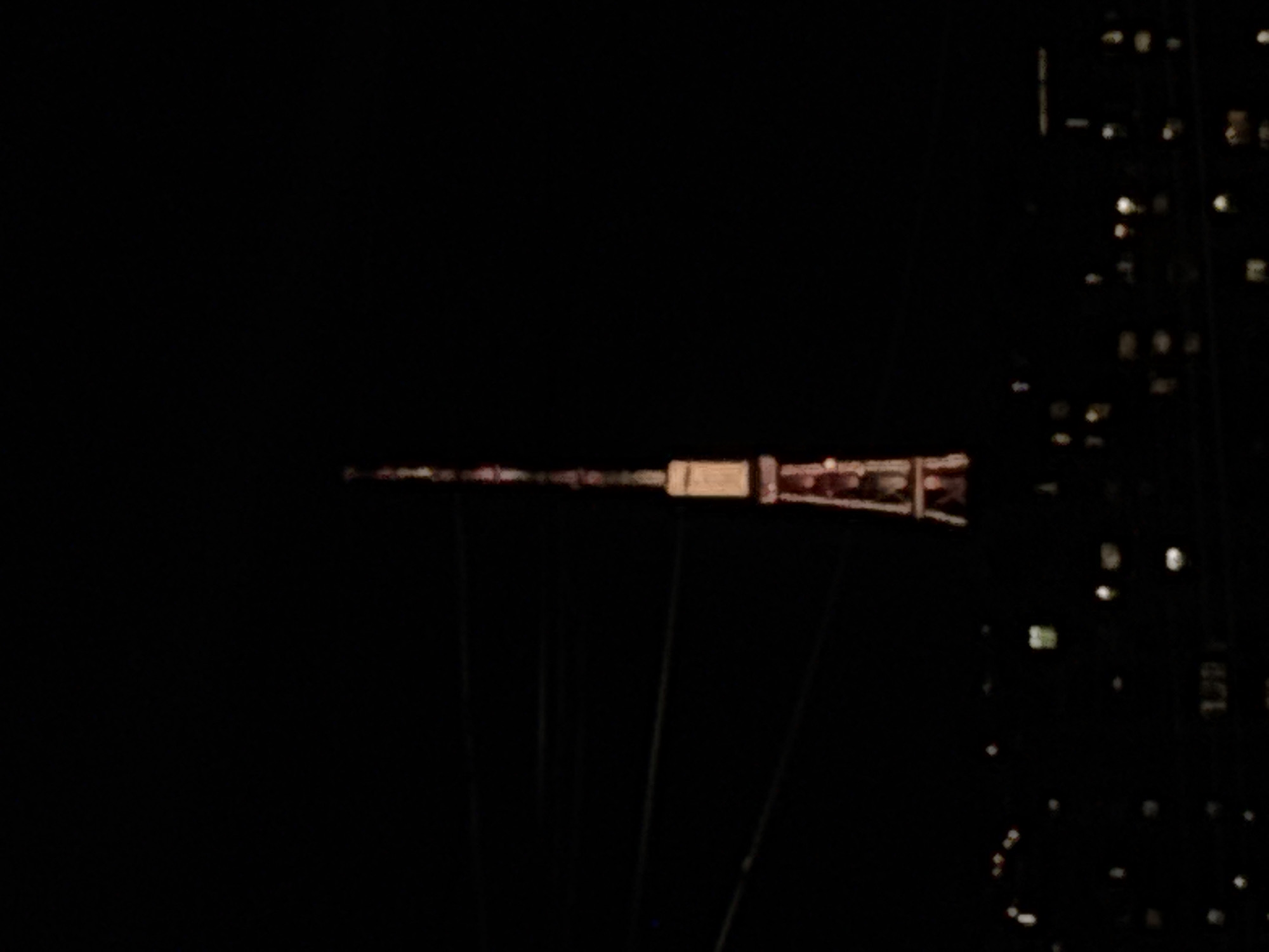 The image displays a night scene dominated by deep blackness, indicative of an urban environment after dark. Centrally positioned and horizontally oriented, a long, slender, illuminated structure pierces the darkness. This structure, likely a tower or a spire, features several glowing segments along its length, culminating in a more substantial, brightly lit section towards its right end, which appears to be enclosed or has a more intricate design.
Below this main horizontal structure, several faint, dark, vertical lines are subtly visible, possibly representing cables or supporting elements extending downwards. To the right of the frame, a vertical section of a building is apparent, distinguished by an array of small, illuminated windows scattered across its dark facade, reinforcing the impression of a city at night.
The overall impression is one of stillness and solitude, with artificial lights providing the only points of interest against the dark backdrop. There is no discernible activity, specific weather conditions, or emotional content conveyed. No visible text can be identified. Based on the provided location context, the prominent illuminated structure is likely the Yerevan TV Tower in Yerevan, Armenia.