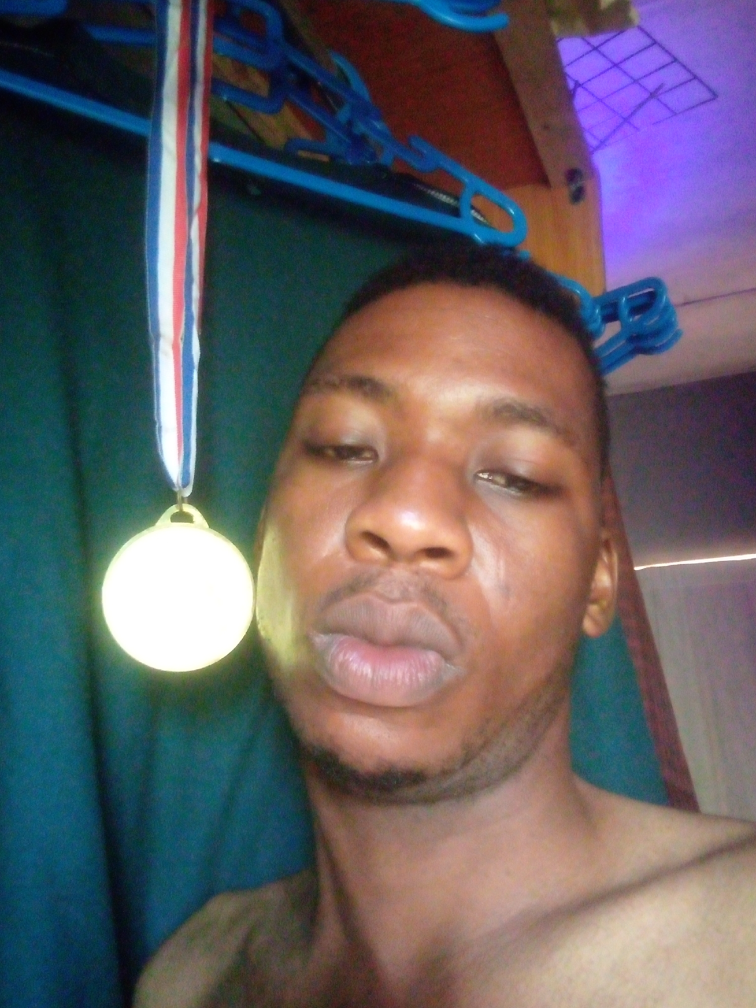 A selfie of a bare-chested Black man is presented with a gold-colored medal on a red, white, and blue ribbon around his neck. The background consists of clothes hangers and a turquoise curtain. The location is unknown; the request for Ikorodu, Nigeria is not reflected in the image.