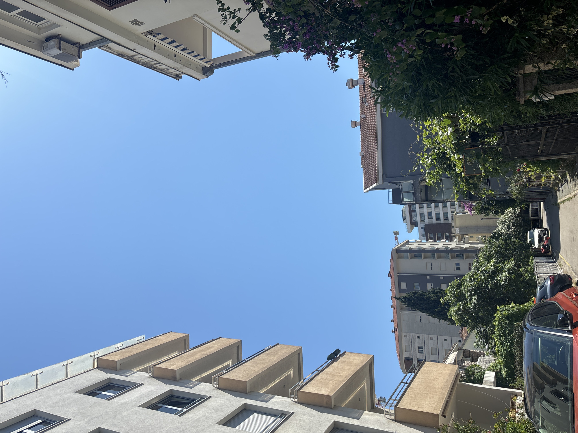 An outdoor, low-angle, eye-level shot captures a street in Budva, Montenegro, under a clear blue sky. On the left, the exterior of a modern apartment building with light grey walls, white trim, and multiple balconies with brown railings and beige facades is visible. On the right, another apartment building with white walls and a blue and white striped awning above a balcony is partly visible, as is lush green foliage with pink flowers. Further down the street, more apartment buildings and trees can be seen. A red car is parked on the left side of the street, and a white car is parked further down on the right, with two figures visible near it. The scene appears to be a residential area with a mix of older and newer architecture, typical of many Balkan coastal towns.