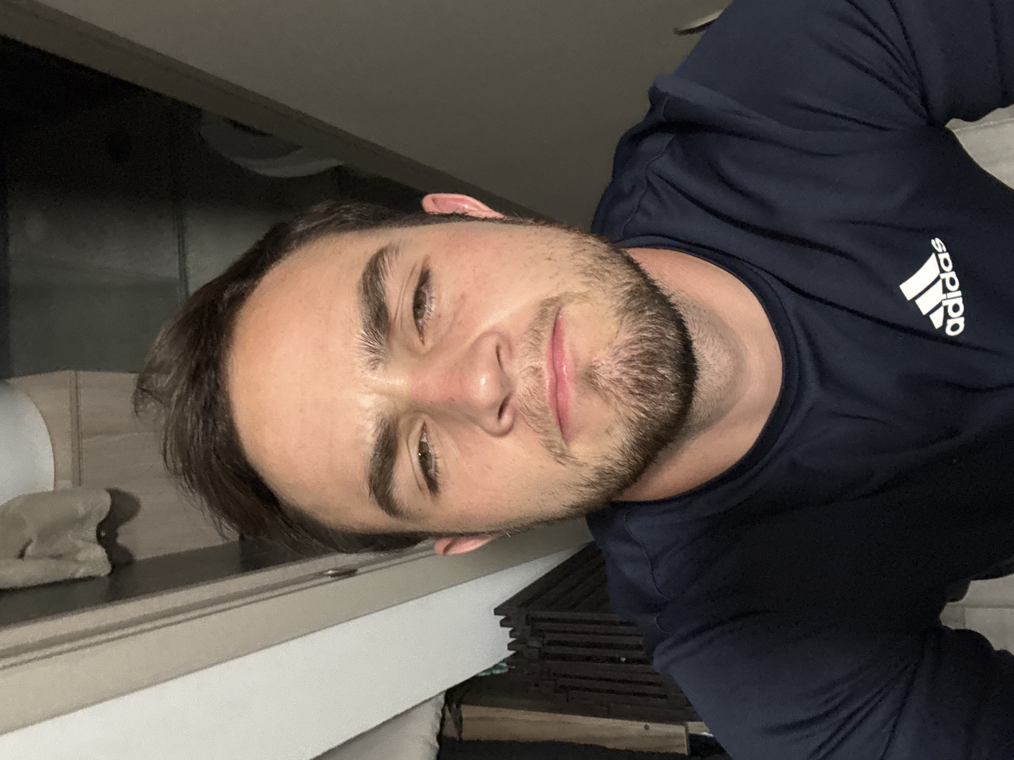 A close-up, rotated portrait features a young man with short dark hair, stubble or a short beard, and dark eyes, looking directly at the camera with a relaxed, slightly smirking expression. He is wearing a navy blue t-shirt with the white Adidas logo, comprising the "adidas" text and the three-stripe emblem, prominently displayed on the right side of his chest.
The scene is set indoors, likely in San Bernardino Tlaxcalancingo, Mexico, under dim artificial lighting. In the background, to the man's left, a light-colored object, possibly a folded towel or cloth, rests on a dark-surfaced counter or shelf. Below and behind him, dark, stacked, slatted objects, potentially made of wood, are partially visible. The image is composed from a low-angle perspective, focusing on the man's face and upper body, conveying a casual, selfie-like moment.