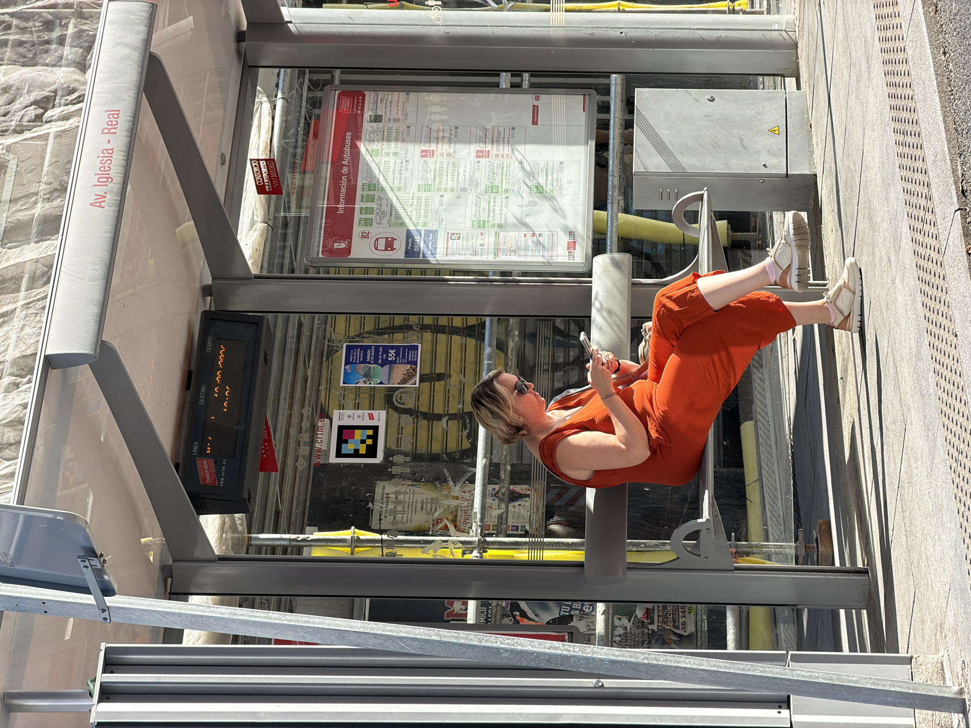 A medium shot captures a woman with blonde hair, wearing sunglasses and an orange jumpsuit, sitting on a bench at a bus stop. She is looking down at her smartphone. The bus stop has a transparent roof and metal supports. Behind her, there is a large timetable displaying bus information. The location is identified as Av. Iglesia - Real in Las Rozas de Madrid, Spain, as indicated by the signage at the bus stop. The ground is paved with grey tiles and a textured pattern. The overall environment suggests an urban setting on a sunny day, with scaffolding visible in the background.