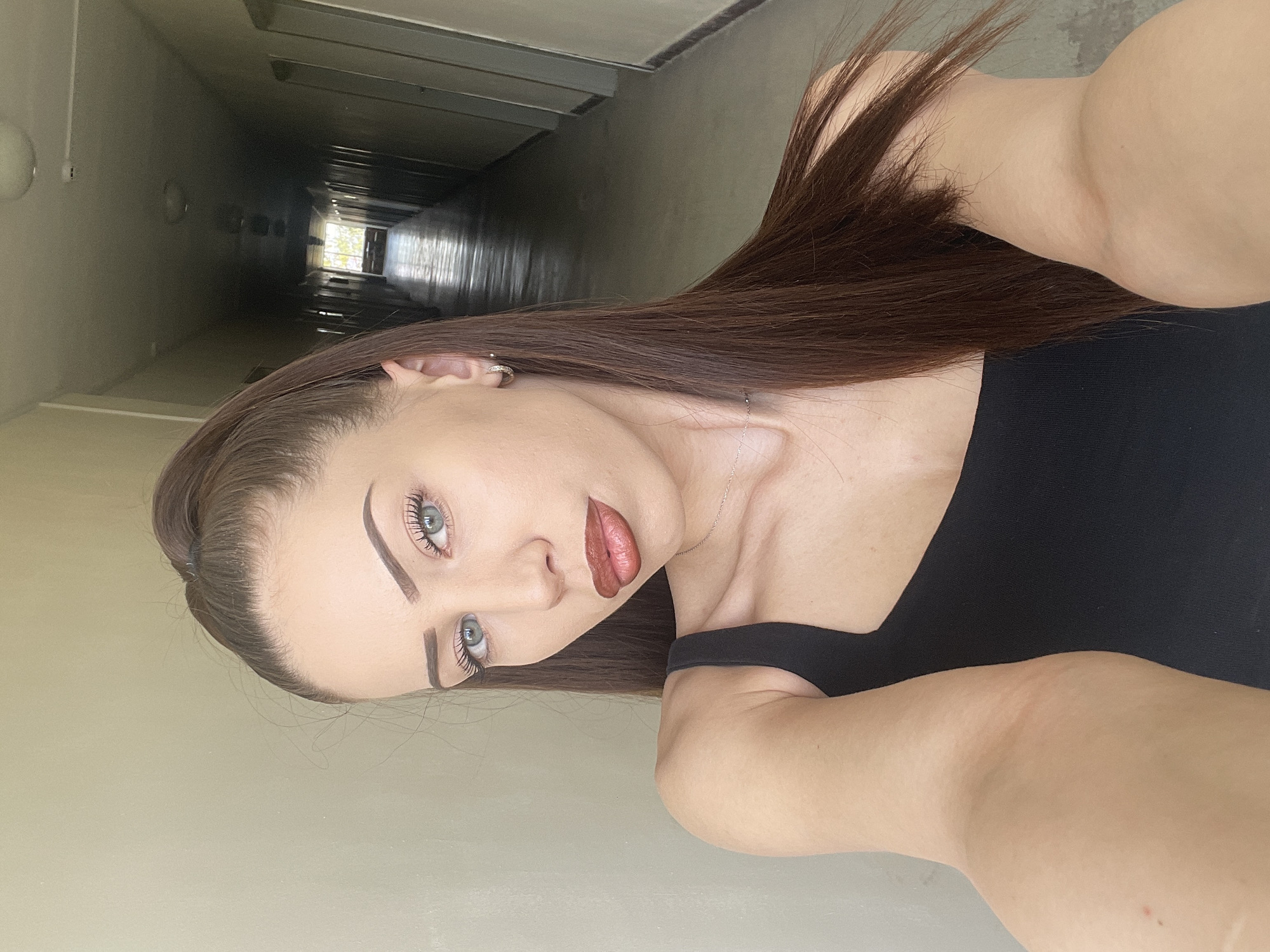 The image captures a young woman posing for a selfie in a long, dimly lit hallway. Her long, straight dark brown hair falls over her shoulder. She has striking blue-green eyes, enhanced with dark eyeliner and mascara, and well-defined eyebrows. Her lips are painted with a reddish-brown lipstick. She wears a black, sleeveless top and a delicate silver necklace. A small, silver hoop earring is visible in her left ear. Her skin is fair, and her expression is neutral.
The background features a deep, narrow corridor with light-colored walls, appearing light grey or beige. The hallway recedes into shadows, with a distant bright light source, likely a window, at its far end, illuminating a section of the floor and casting some reflections. The overall ambiance of the hallway is stark and utilitarian. The subject is brightly lit, creating a strong contrast with the shadowed background. This scene is located in Veliko Tarnovo, Bulgaria. No visible text is present.