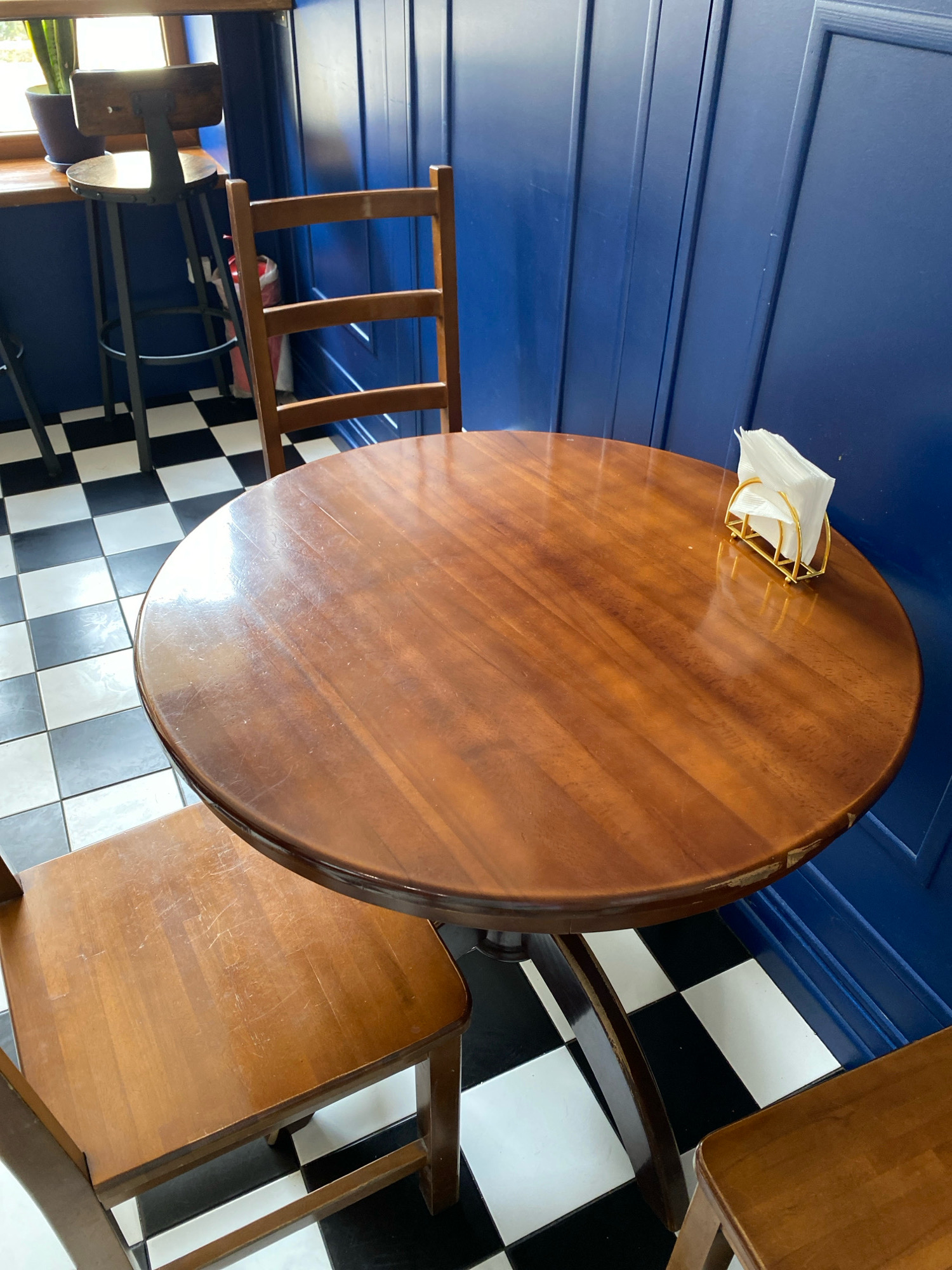 This is a medium shot of a round, wooden table in a cafe setting in Chornomorsk, Ukraine. The table is made of polished wood with visible grain and some surface scratches. A gold napkin holder with a stack of white napkins sits on the right side of the table. Two wooden chairs are partially visible. One chair is to the left of the table, and the seat is in focus. Another chair is to the right, with only the seat and part of the leg visible. The floor is a black and white checkered tile pattern. In the background, a blue paneled wall runs vertically. To the far left, a sliver of a window and a portion of a tall bar stool with a wooden seat and metal legs are visible. A potted plant is on the windowsill. The overall impression is that of a clean, simple cafe interior.