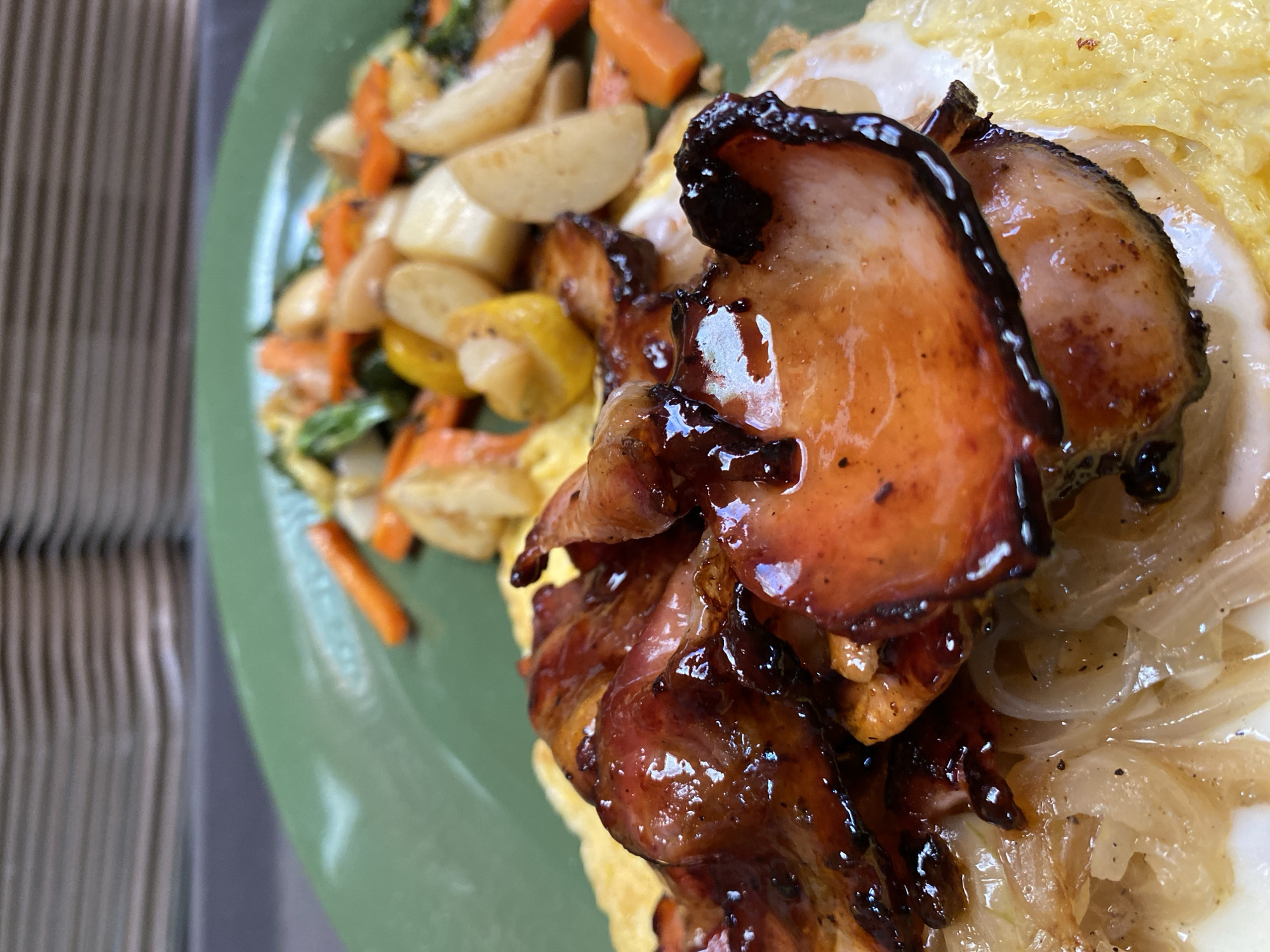 A close-up, slightly tilted shot shows a plate of food. On the left side of the green plate is a mix of stir-fried vegetables including carrot sticks, yellow bell pepper slices, potato chunks, and green vegetables. The right side has a dish that appears to have a base of a yellow omelet or scrambled egg. On top of the egg is a layer of sauteed onions, and the main element is a pile of glazed ham pieces. The glaze on the ham is dark brown. No people are visible. The scene does not provide sufficient clues to pinpoint the location to Puebla, Mexico.