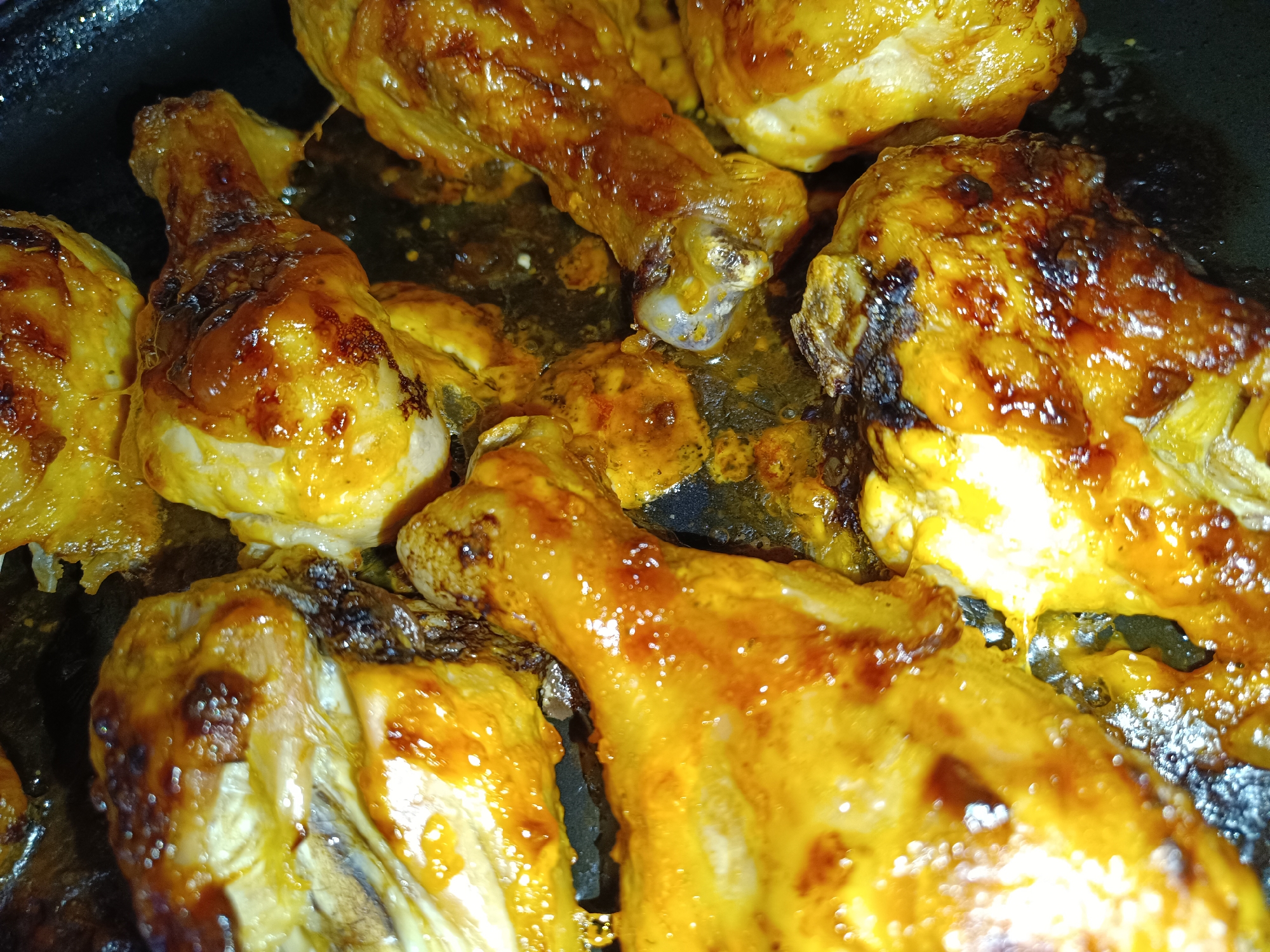 The image shows a close-up of cooked chicken pieces in a dark pan. The chicken appears to be glazed and has a golden-brown color. Some pieces show darker, slightly charred areas. There's some rendered fat or sauce at the bottom of the pan. The scene shows only the chicken and the pan and no evidence of where this photo was taken. It is not possible to tell if the image was taken in Tobolsk, Russia, or any other location.
