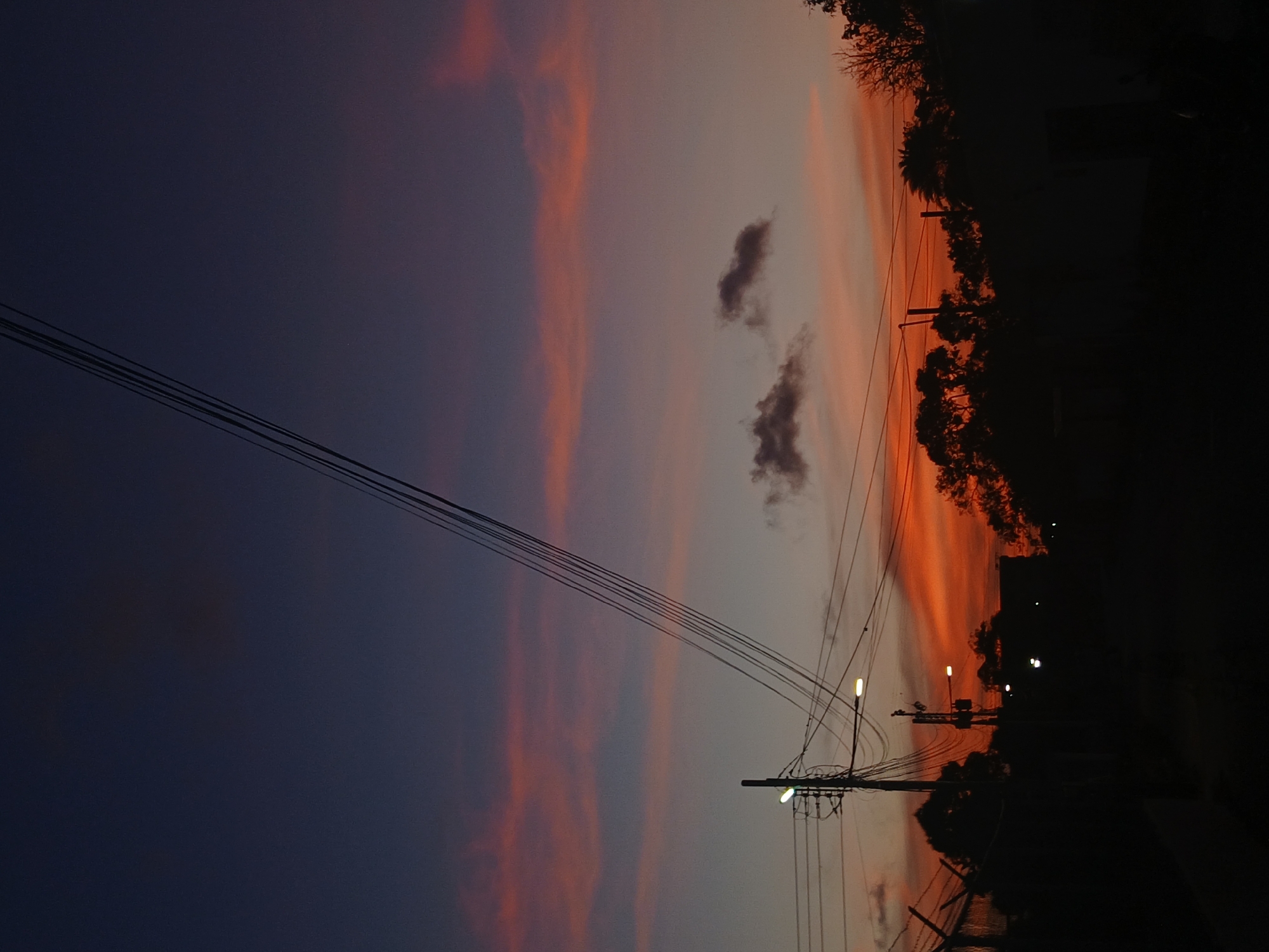 This image, rotated 90 degrees clockwise to be viewed upright, captures a dramatic sunset scene, likely over an urban or suburban landscape in Juan Griego, Venezuela.
The sky dominates the upper two-thirds of the frame, displaying a striking gradient of twilight colors. It transitions from a deep, dark indigo and purple at the top to softer purples, vibrant pinks, and fiery oranges and reds closer to the horizon. Long, streaky clouds are visible, some reflecting the intense sunset hues, while two distinct, darker cloud formations appear in the lighter parts of the sky.
In the mid-ground, a prominent utility pole stands, supporting a complex network of overhead power lines that stretch across the frame and into the distance. Several streetlights are attached to this pole and other poles further along, casting warm, yellow-orange light against the deepening dusk.
The lower third of the image is rendered in a dark silhouette against the bright horizon. This includes a dense line of trees and bushes, possibly containing palm trees, as well as indistinct shapes that could be buildings or parts of a fence. The ground or a road surface is also visible in deep shadow. No people or visible text are present. The illuminated streetlights confirm the deepening twilight, creating a serene and atmospheric tableau.