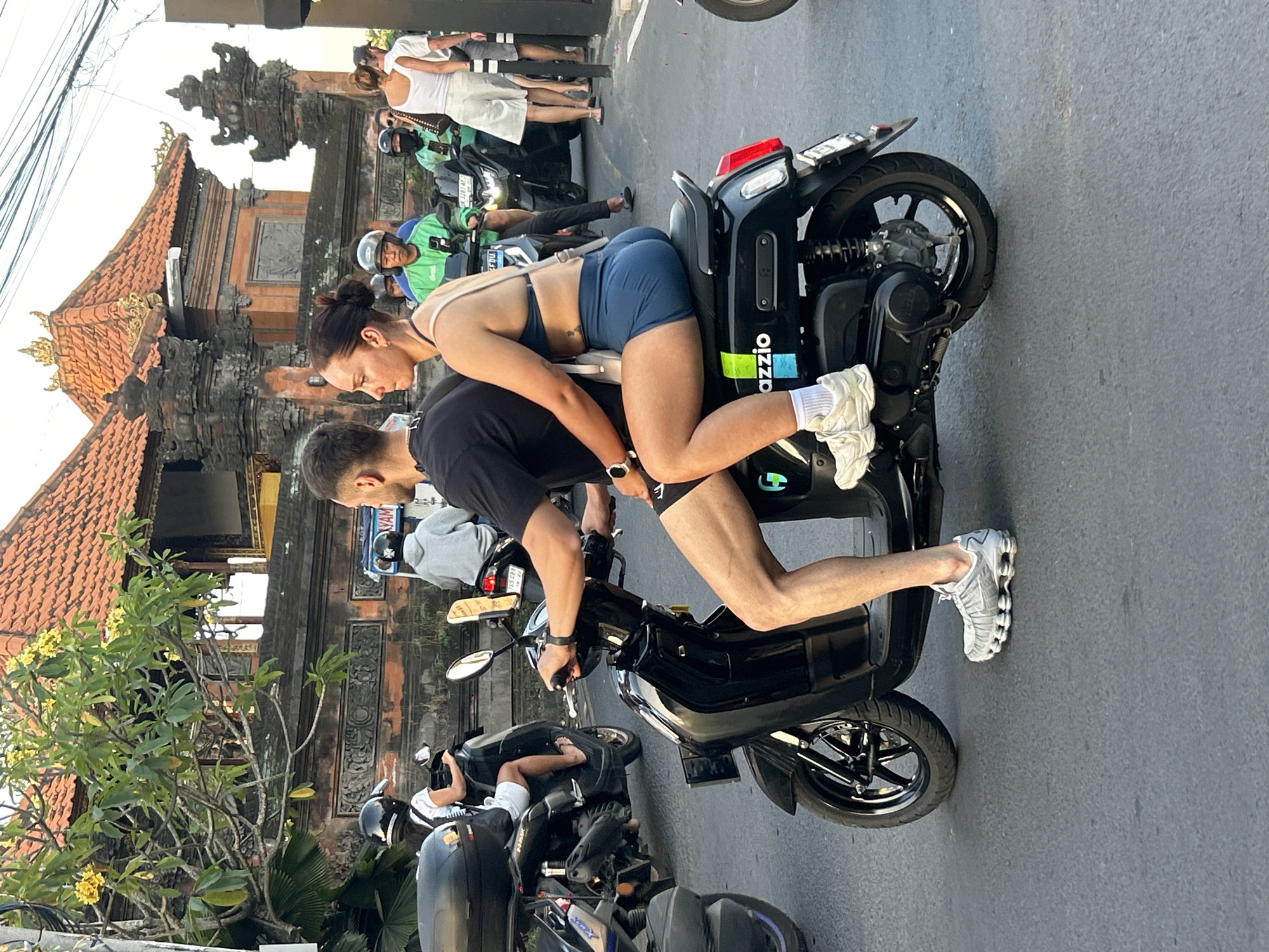 In North Kuta, Indonesia, on a paved street, a man and woman ride a black electric scooter. The man drives, wearing a black t-shirt and dark shorts, with gray athletic shoes. The woman is the passenger, dressed in a blue sports bra and blue shorts, with white athletic shoes and white socks. She has her arm around the man. The scooter has the brand "gOzzio" visible on its side. In the background, other scooters and people are present. A building with traditional Balinese architecture, featuring terracotta roofs and ornate stone carvings, is visible behind them. The lighting suggests it is daytime, possibly late afternoon, with some sunlight casting shadows.