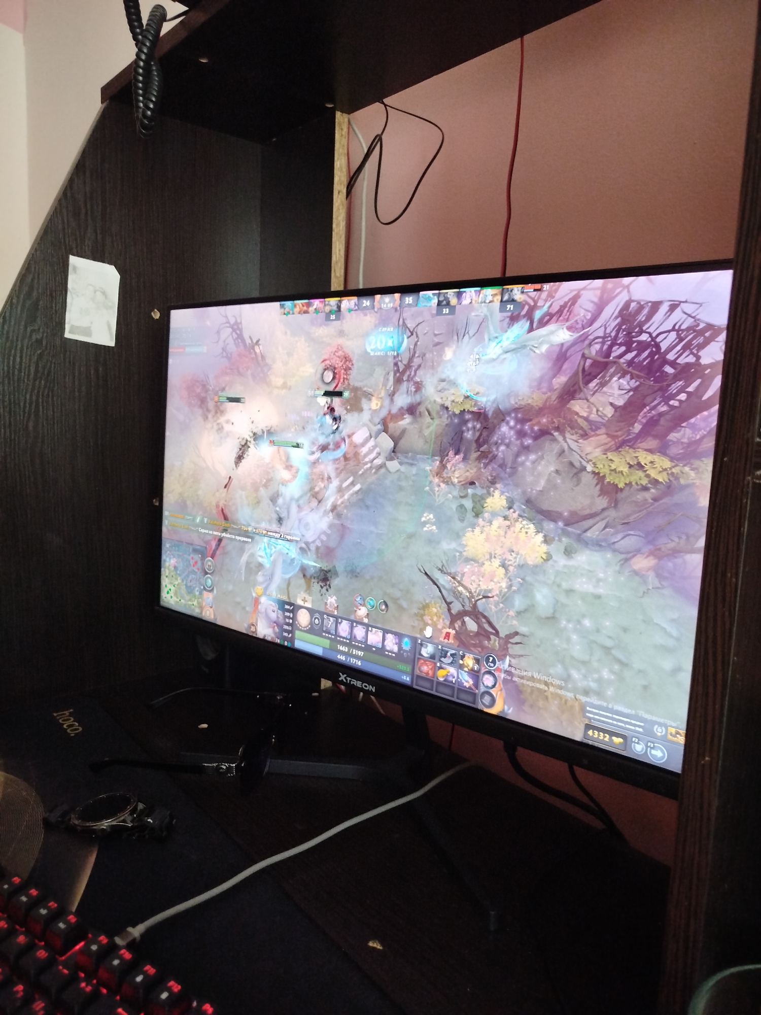 This media file shows a close-up of a computer monitor displaying a video game. The game appears to be a real-time strategy or multiplayer online battle arena (MOBA) game, with various characters and map elements visible on the screen. The game's interface shows player statistics, health bars, and item icons.
In the foreground, a portion of a keyboard with red backlighting is visible, suggesting a gaming setup. A dark-colored mousepad with the "hoco." logo is also present, along with a smartwatch and a pair of glasses. The monitor is set within a dark wooden enclosure, possibly a gaming booth or a desk with built-in shelving. Cables are visible, including a black coiled cable hanging from above and a white USB cable.
The scene is dimly lit, indicating the focus is on the monitor's display. The overall impression is one of a dedicated gaming environment. The text visible on the monitor includes game-related information and a prompt to activate Windows. The game itself features a fantasy or sci-fi aesthetic with vibrant colors and detailed graphics. No people are directly visible in the image, but the setup implies a player is present.