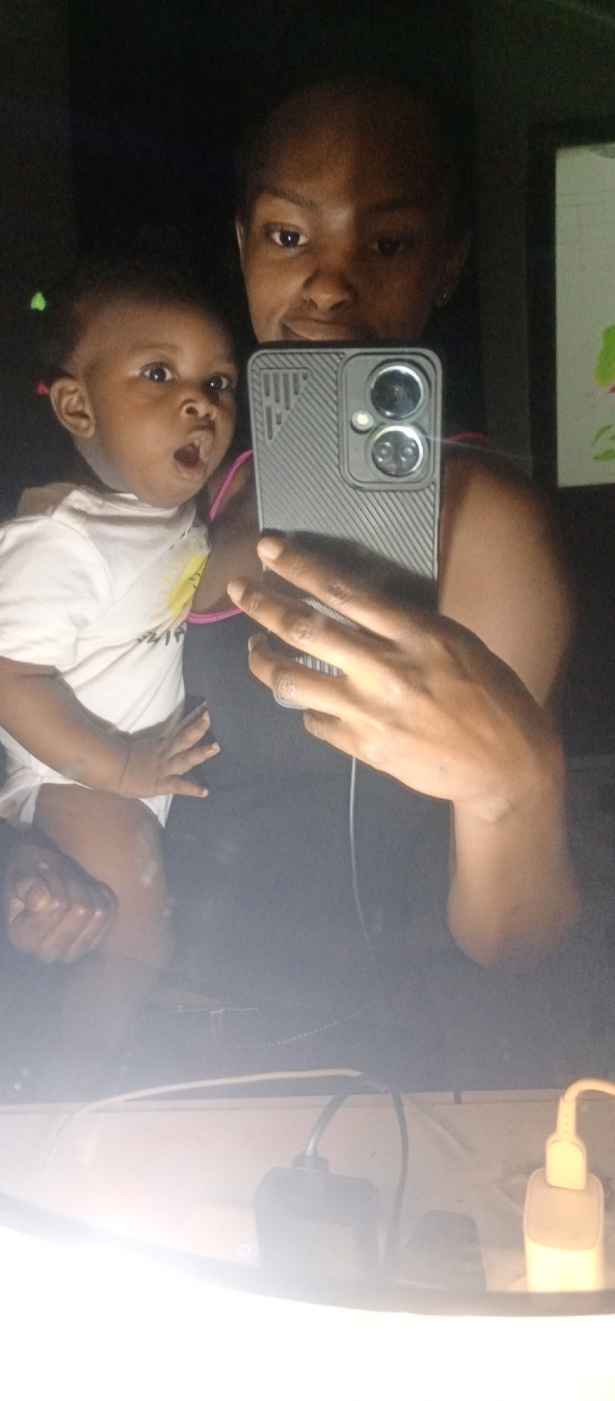 A person is holding a young child while taking a selfie in a dimly lit room. The person is holding a phone with two prominent camera lenses and a textured gray case. The child is wearing a light-colored shirt and has an expression of surprise or astonishment. In the background, there is a partial view of a wall with some artwork. The lower portion of the image shows a surface with electrical outlets and charging adaptors. The setting is unknown.