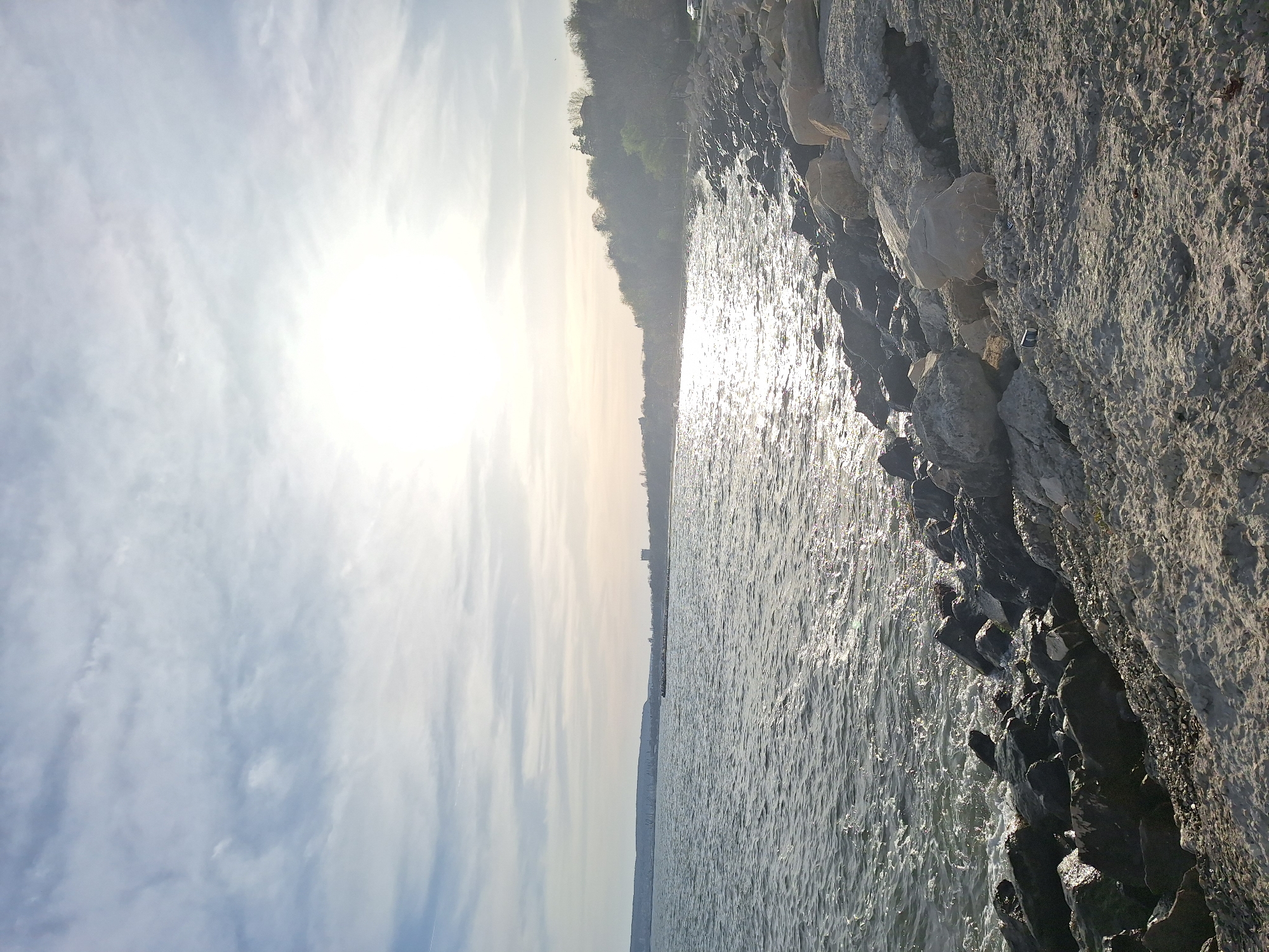 The image shows a waterfront scene in Varna, Bulgaria. The sun is bright and slightly obscured by clouds in the upper left. The Varna coastline stretches across the horizontal center, with buildings barely visible on the horizon. The water shimmers with sunlight. A rocky breakwater extends along the right side of the image.