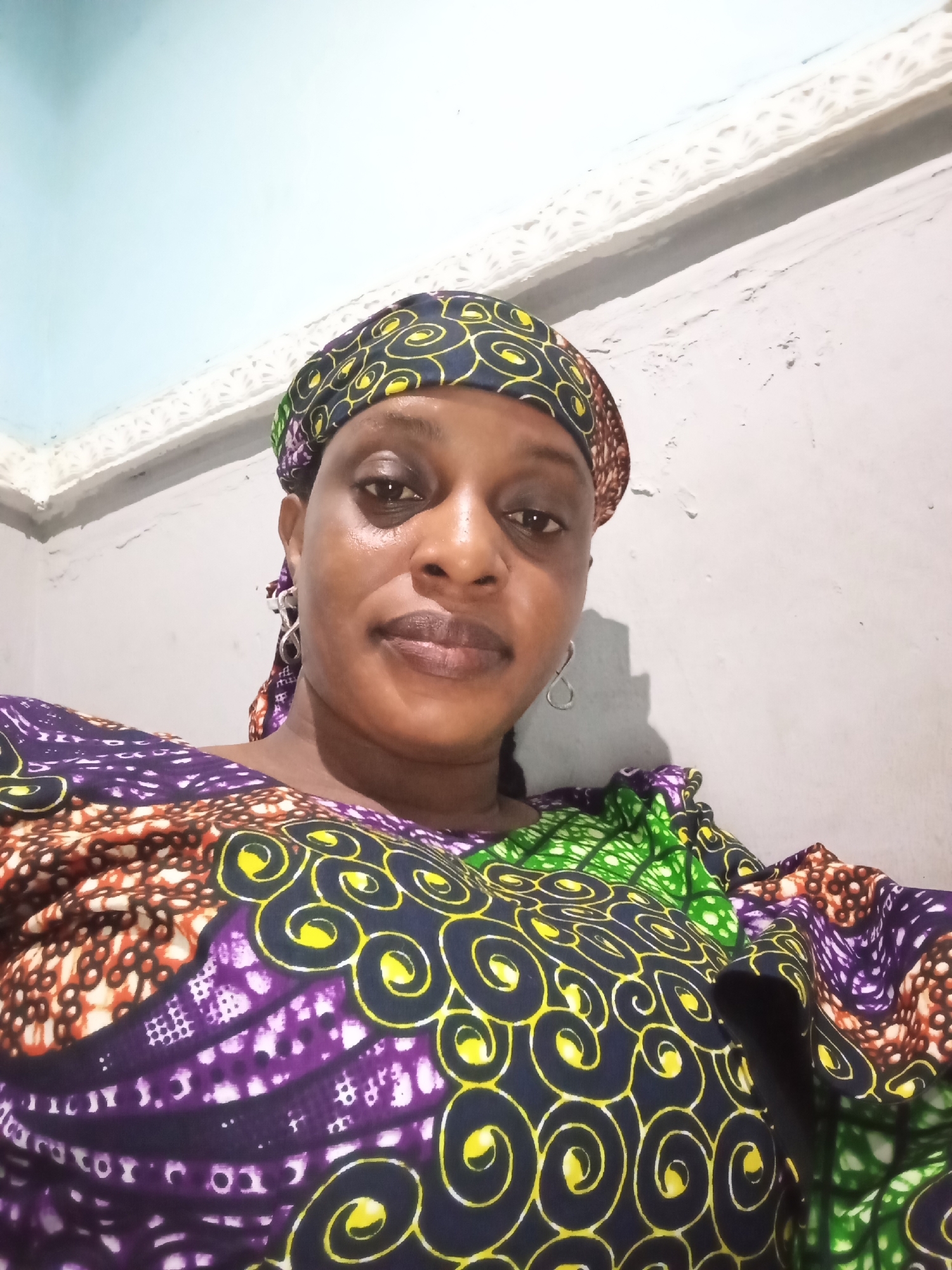 A woman is facing the camera. She wears a colorful headscarf and a dress with similar patterns. Her earrings are visible. The background is a wall with patchy paint. The location is Unknown, Unknown.