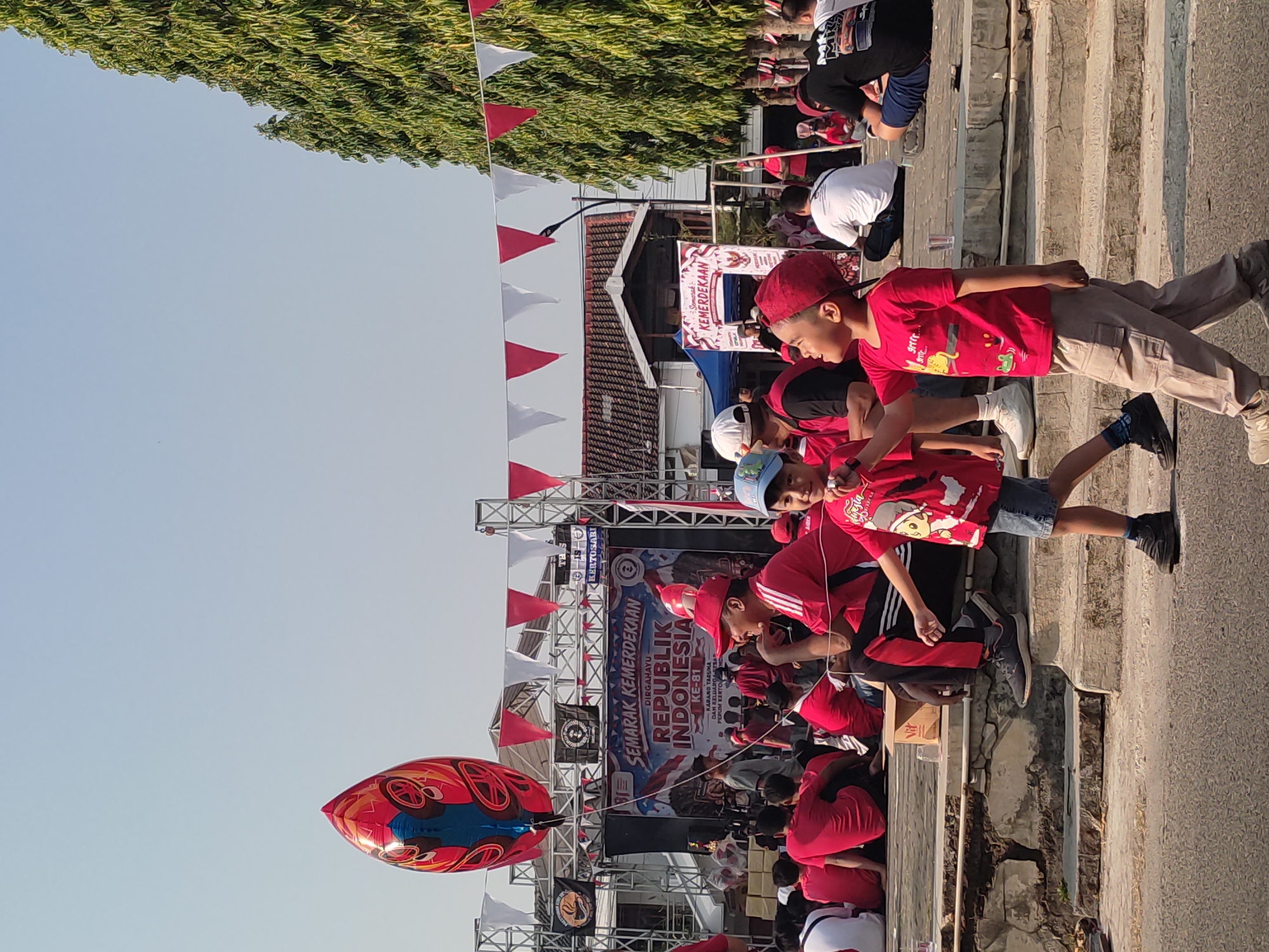 The image depicts an outdoor event, likely a celebration, in Ponorogo, Indonesia. A large, brightly colored balloon, predominantly red and blue with yellow accents and a design resembling a stylized face or mask, floats in the sky. In the background, a stage is set up with banners that read "SEMARAK KEMERDEKAAN REPUBLIK INDONESIA KE-81" (Grand Celebration of the 81st Anniversary of the Republic of Indonesia). Red and white triangular flags are strung across the area, common decorations for Indonesian Independence Day.
Numerous people are present, many wearing red shirts, suggesting a unified group or theme. In the foreground, two young boys are prominent. The boy on the left, wearing a red shirt and a blue cap, is holding onto a string. The boy on the right, also in a red shirt and a red cap, is walking and looking upwards, possibly at the balloon. Several adults are seated in the middle ground, observing the scene. One man in the foreground, wearing a red shirt and a red cap, is looking to his right and smiling. The environment appears to be a public space with trees and buildings visible in the background. The ground is paved, and there are tiered stone structures in the middle ground where people are seated. The sky is clear and bright, indicating a daytime event. The overall atmosphere seems festive and communal.