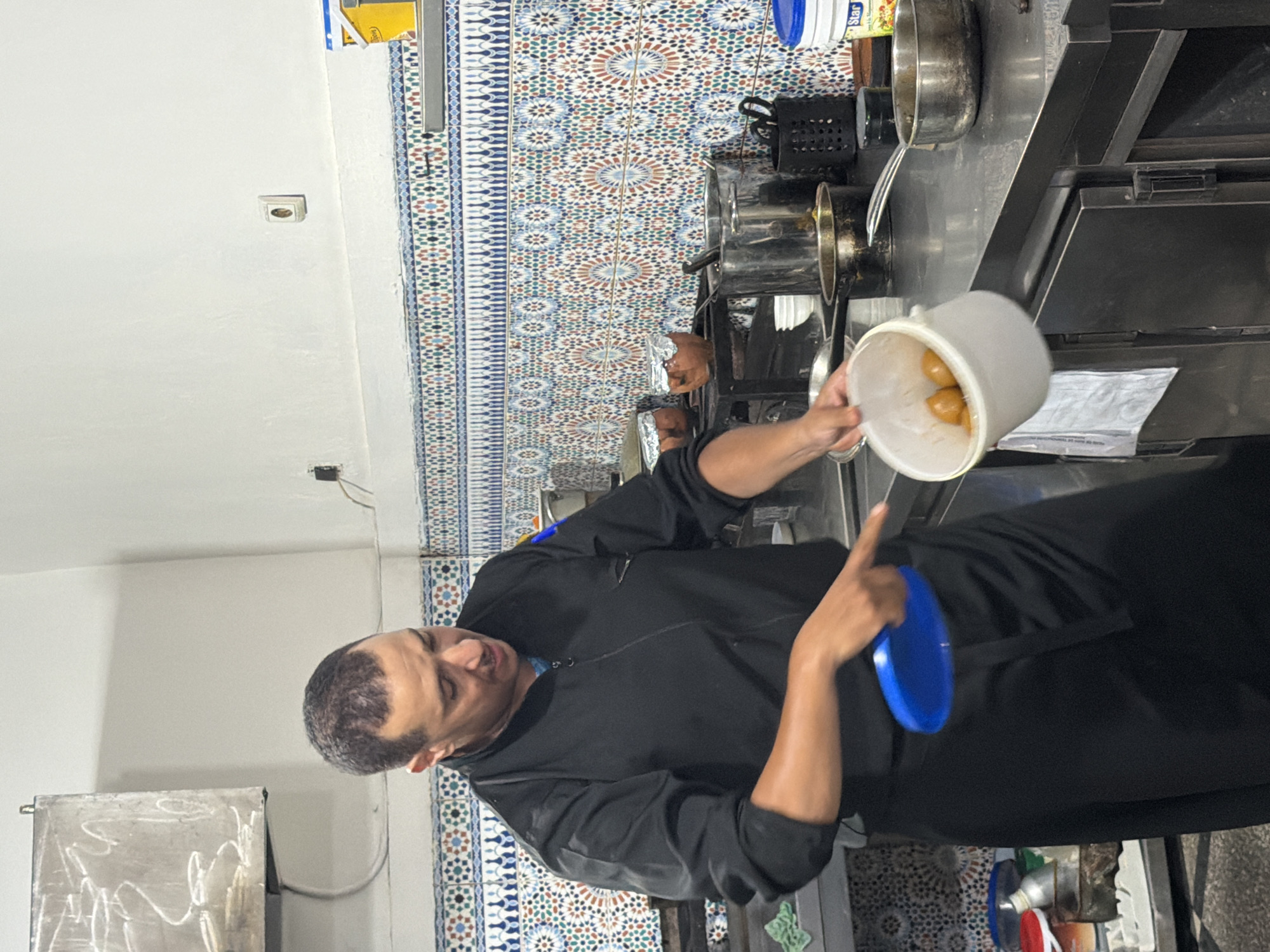 A man wearing a black chef's jacket and apron is standing in a kitchen, likely in Marrakesh, Morocco, given the decorative tile backsplash. He is holding a white bucket containing some round, brown food items, possibly pastries or dumplings, and appears to be in the process of emptying them or preparing them for cooking. His left hand is extended, holding a blue plate, while his right hand is near the bucket. The kitchen counter is made of stainless steel and is covered with various cooking implements, including pots, pans, and a utensil holder. Behind the man, the wall is adorned with intricate mosaic tiles in shades of blue, red, green, and white, a common feature in Moroccan architecture. The overall scene suggests a busy culinary environment where food preparation is underway.