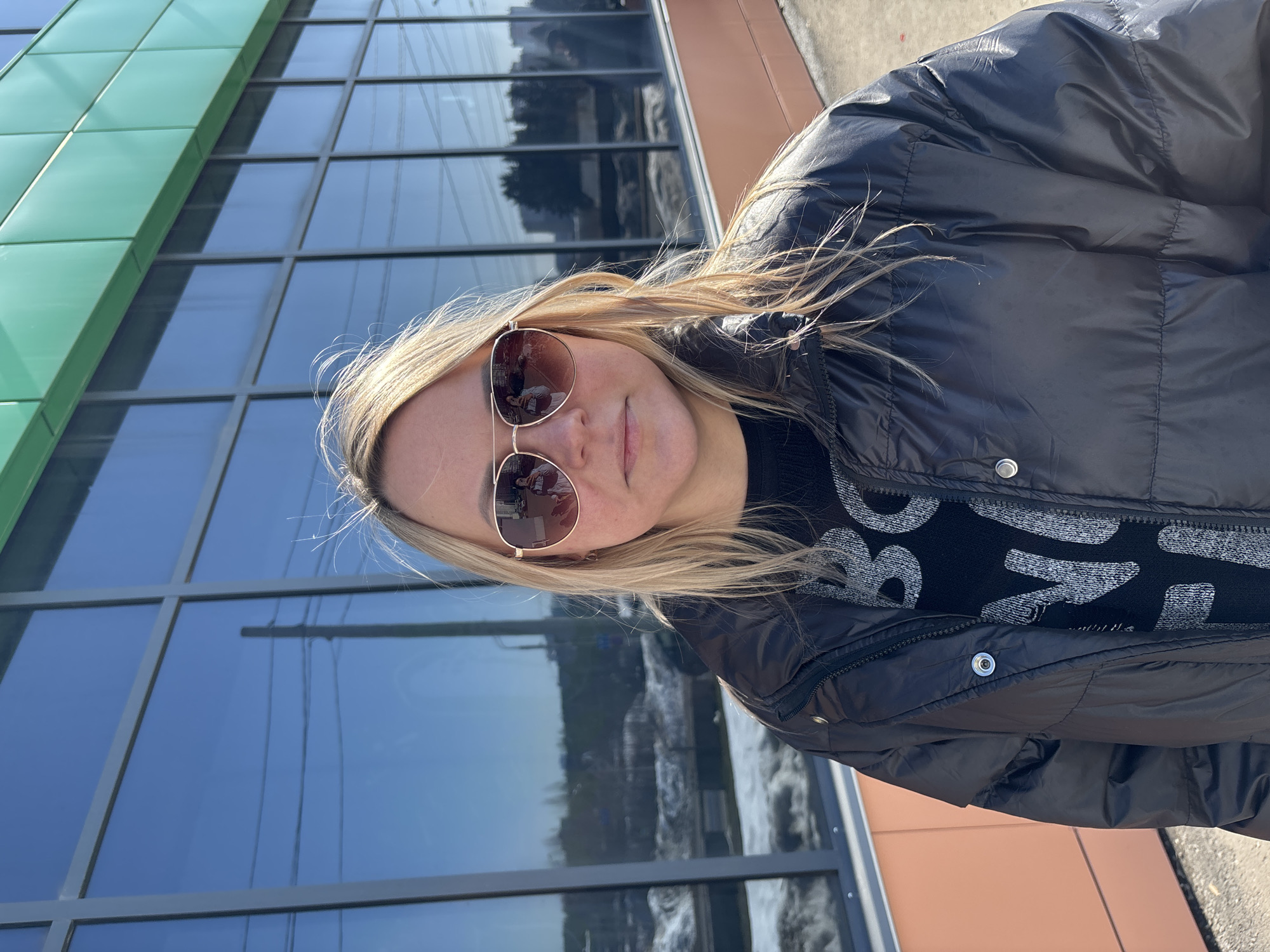 A single individual, identified as female, stands upright and faces the camera directly. She has blonde hair and wears sunglasses with gold-toned frames and brown-tinted lenses. Her attire consists of a black, possibly nylon, puffer jacket, partially unzipped, revealing a black knitted sweater with light-colored, patterned lettering. Her facial expression is neutral.
The scene is outdoors, on a paved surface, in front of a commercial building identified as the "Omega" Shopping Mall (ТРЦ Омега) located at K. Marx St, 201, Izhevsk, Russia. The building's facade features a lower section clad in orange-peach panels, a middle section composed of large, dark-tinted, reflective glass windows within a grey grid frame, and an upper section with green facade panels.
The environmental conditions indicate a clear, sunny day, with strong sunlight casting reflections on the building's windows and the subject's eyewear. Patches of snow are visible on the ground in the background, suggesting a cold-weather season. Reflections in the glass surfaces reveal elements of an urban environment, including power lines, utility poles, and distant buildings.