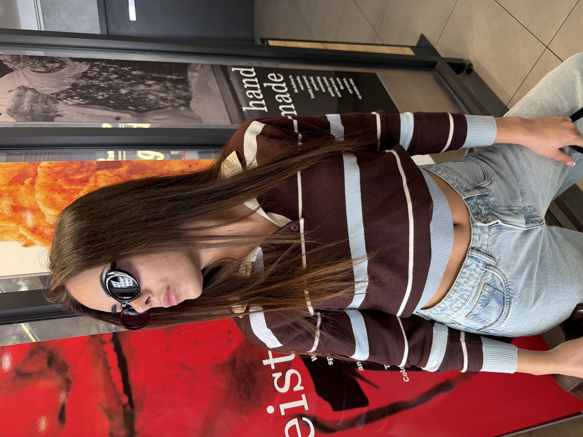 The image features a woman wearing sunglasses, a brown and blue striped top, and light blue jeans, positioned indoors. She is in front of a vertical food advertisement. The advertisement has a red background with the white text "eist" vertically printed. Above it, there are images related to food preparation and presentation, including one that says "hand made". The environment appears to be a restaurant or food establishment. The location is likely in Sofia, Bulgaria, but there is no definite information about its location in the city.