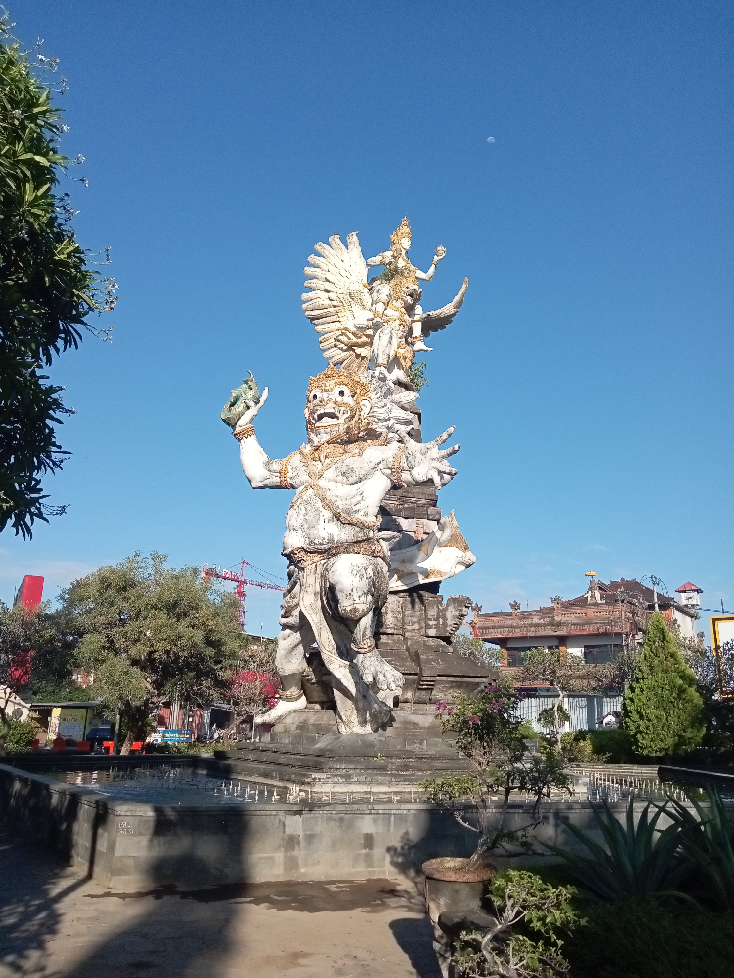 This image, taken in the city of Unknown, Unknown, depicts a large, weathered multi-component stone statue under a clear, vibrant blue sky with a faint crescent moon visible in the upper right. The primary figure at the statue's base is a colossal, white, humanoid entity with a fierce, fanged expression, large eyes, and an elaborate gold crown and body ornamentation. This figure is depicted in a squatting or kneeling posture, holding a green, urn-like object in its raised left hand. Its right hand is extended. The figure shows signs of age and discoloration.
Stacked above and slightly behind this main figure is a smaller, golden-hued deity riding on the back of a large, white-winged bird, identified as Garuda, with its wings spread wide. This deity also wears a crown and holds an object in its hand. Both parts of the statue rest on a multi-tiered dark gray stone pedestal.
In the foreground, a dark gray, multi-level fountain or pool surrounds the statue's base, with visible water jets. The words "BALI SARI" are carved into the side of the fountain. The paved ground around the fountain shows distinct shadows, indicating strong sunlight. Various potted plants, including a bonsai-style tree, and bushes are arranged around the fountain structure.
To the left of the statue, the dense green foliage of a large tree with small white flowers extends into the frame. In the mid-ground and background, several buildings with traditional architectural features, including multi-tiered brown roofs and ornate decorative elements, are visible. One building features a distinctive yellow umbrella on its roof. A large red construction crane is visible above the trees in the upper left background.