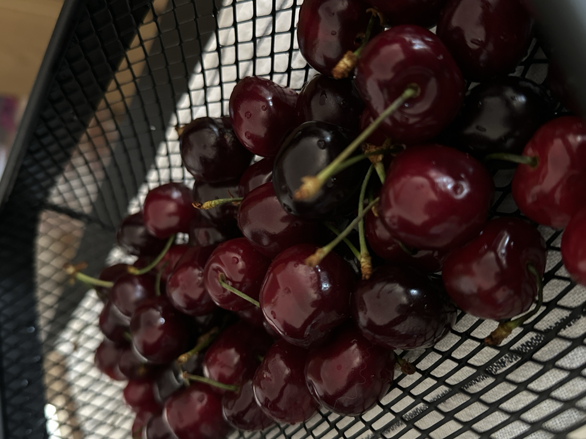 A close-up, slightly angled shot shows a black wire basket filled with ripe, red cherries. The cherries have dark red to almost black skin, with some displaying small water droplets. Green stems protrude from the tops of several cherries. The basket is positioned against a blurred background, but the scene's exact location cannot be determined from the image alone. The city and country information are extraneous and cannot be verified.