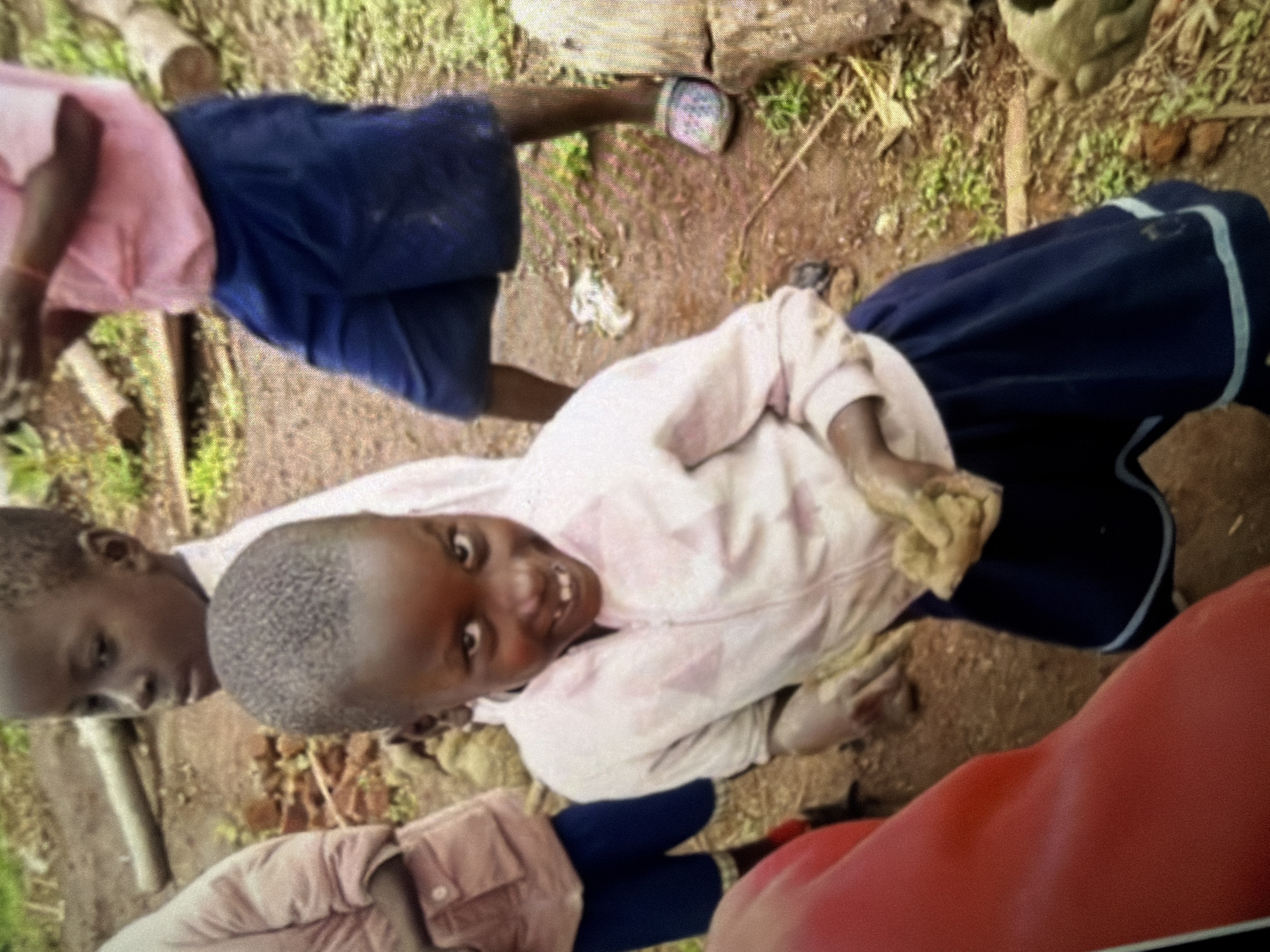 The image shows three children in an outdoor, rural setting. In the foreground, a young Black child with short, dark hair is smiling and looking at the camera. They are wearing a pink jacket with a zipper and a navy blue skirt. Their hands appear to be covered in mud. To the left of this child, another child, also Black, is partially visible from the waist up, wearing a pink jacket. Behind the smiling child and to their left, a third Black child is standing, wearing a pink shirt and navy blue shorts. The ground is dirt and some greenery is visible. Logs and a few scattered rocks are also present in the environment. The overall impression is a candid shot of children playing outdoors.