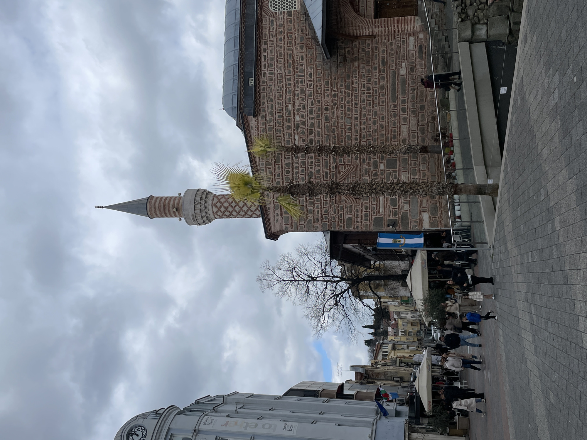An urban pedestrian scene is depicted in central Plovdiv, Bulgaria, featuring the Djumaya Mosque and its surrounding area, including the Ancient Roman Stadium. The Djumaya Mosque, located on the right, presents a facade of reddish-brown brickwork and stone banding, topped by a gray-domed roof. A slender, striped minaret with a conical spire rises prominently beside the mosque. Two mature palm trees stand before the mosque's facade. To the left, a light-colored, multi-story building features a domed clock tower at its apex and displays "Plovdiv 2019" banners. The foreground consists of a grey paved pedestrian square. Approximately two dozen individuals, including adults and several children, traverse this area in various directions, dressed in cool-weather casual attire. Outdoor cafe seating with white umbrellas is visible along the left side of the street. A modern pedestrian bridge with metal railings ascends towards the mosque, spanning a visible excavation of the Ancient Roman Stadium. Two individuals are observed walking upon this bridge, moving towards the mosque. In the background, a bare deciduous tree and a tree-covered hill are visible beneath an overcast sky.