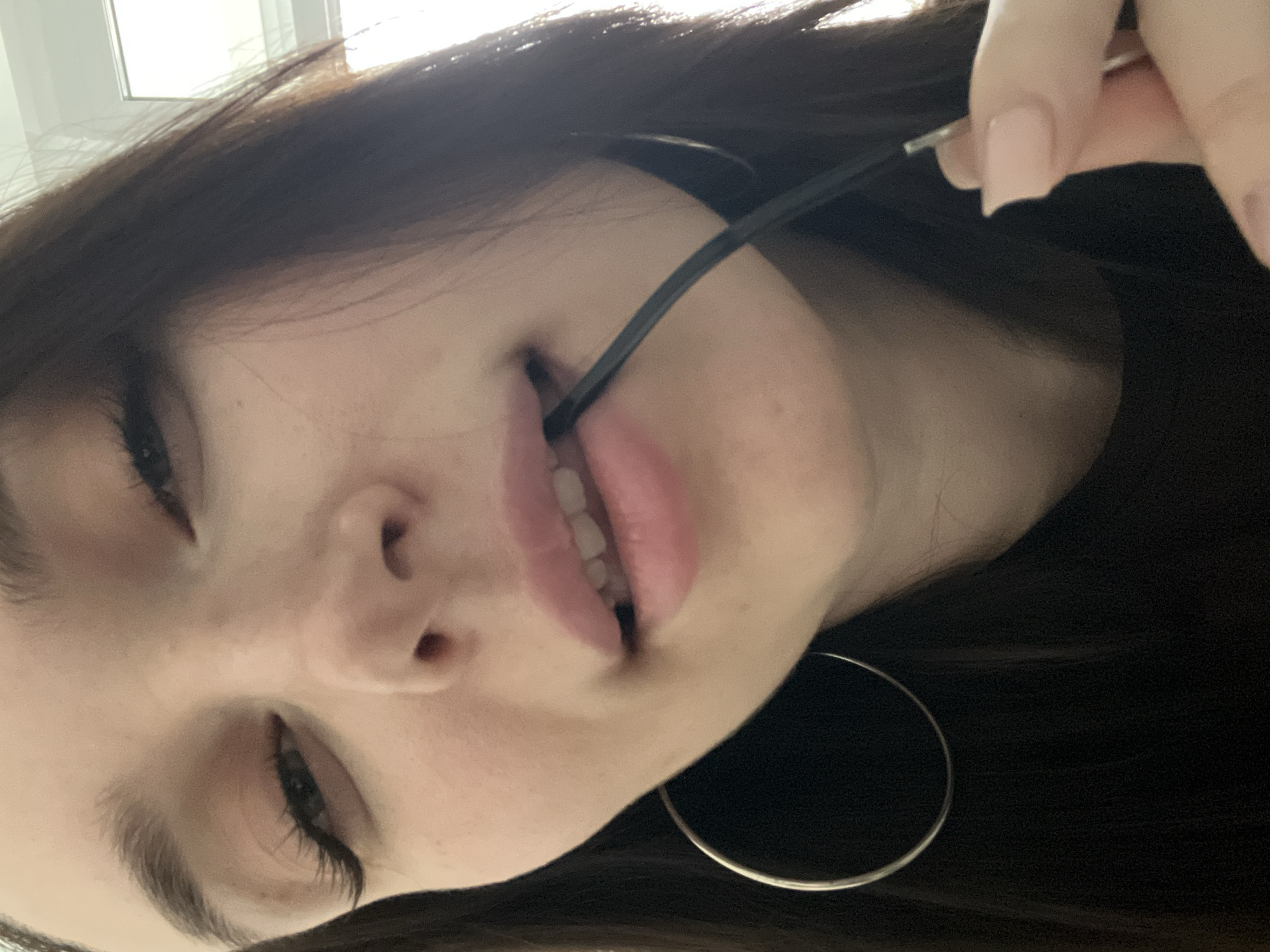 A close-up, angled shot depicts a young woman with long, dark hair. She has light skin, dark eyelashes, and pink lips. A black object, possibly a glasses frame, is held by her right hand with pale pink painted nails and partially held in her mouth. She is wearing a large hoop earring. The background is blurry and indistinct, but a white wall and a light fixture are visible. There is no evidence that this image was taken in the city of Tyumen, Russia.