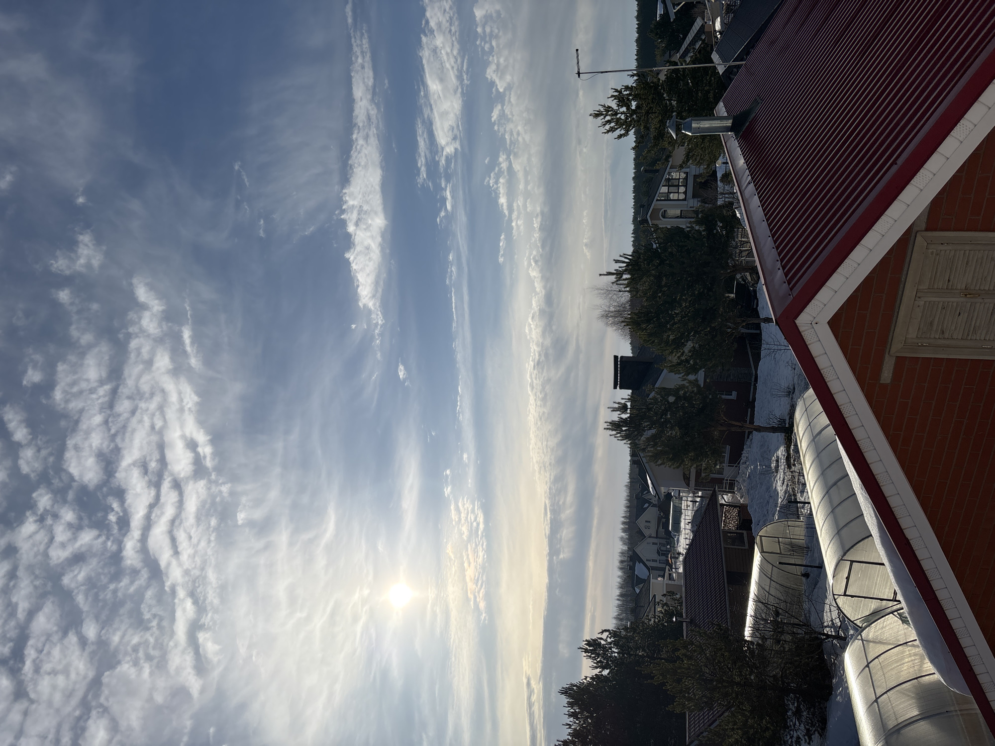 An elevated exterior photograph depicts a residential area in Русский Вожой, Russia, under a bright, partly cloudy winter sky. The sun is a prominent, radiant orb in the upper left quadrant, casting a strong light across the scene.
In the immediate foreground, the upper section of a red brick building is visible on the right, featuring a dormer with light-colored, closed louvered window shutters. Its dark red corrugated metal roof slopes steeply, with a light accumulation of snow along its lower edge. A single, cylindrical metal chimney with a reflective conical cap protrudes from this roof.
The midground reveals a snow-covered landscape dotted with various residential structures and coniferous trees. On the left, three semi-circular, translucent greenhouses or cold frames, with visible internal metal bracing, are aligned on the snow-covered ground, reflecting the sunlight. Beyond these, a cluster of detached single-family houses with diverse architectural styles, including dark grey and brown roofs, are nestled amongst numerous evergreen trees and bare deciduous trees. A pile of split firewood is stacked near one of the houses. A black utility pole with an attached street light fixture is present on the far right.
The background features a dense, dark green forest line stretching horizontally across the distant horizon, demarcating the edge of the developed area. The sky, occupying the upper two-thirds of the image, is predominantly blue with scattered formations of white, wispy cirrus clouds and some denser, more undulating cloud patterns. No individuals or specific actions are discernible within the frame.